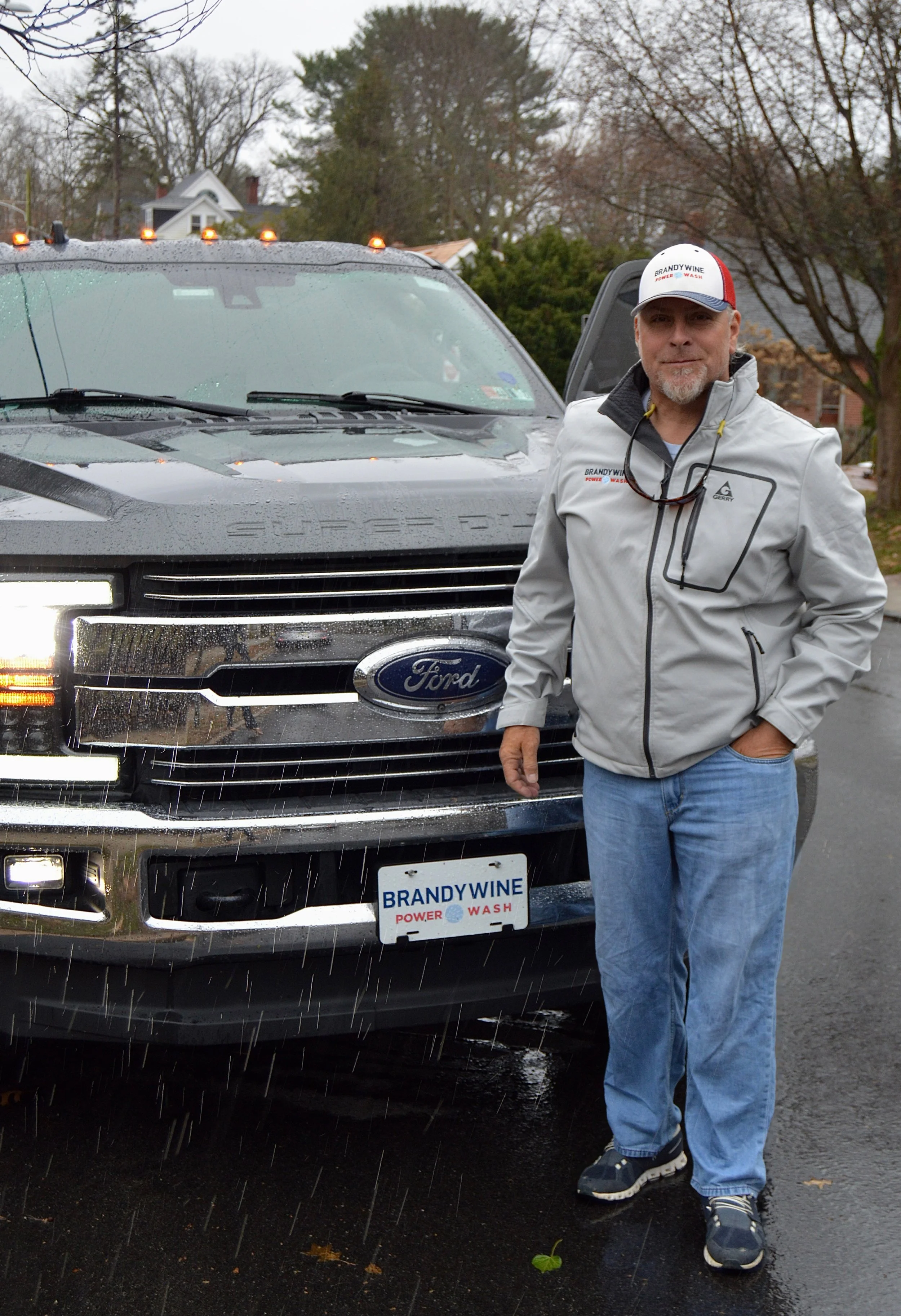 A man standing in front of a black Ford vehicle, wearing a gray jacket, blue jeans, and a white and red cap, with a license plate that reads 'BRANDYWINE POWER WASH'. It is raining, and the man appears to be after a car wash.