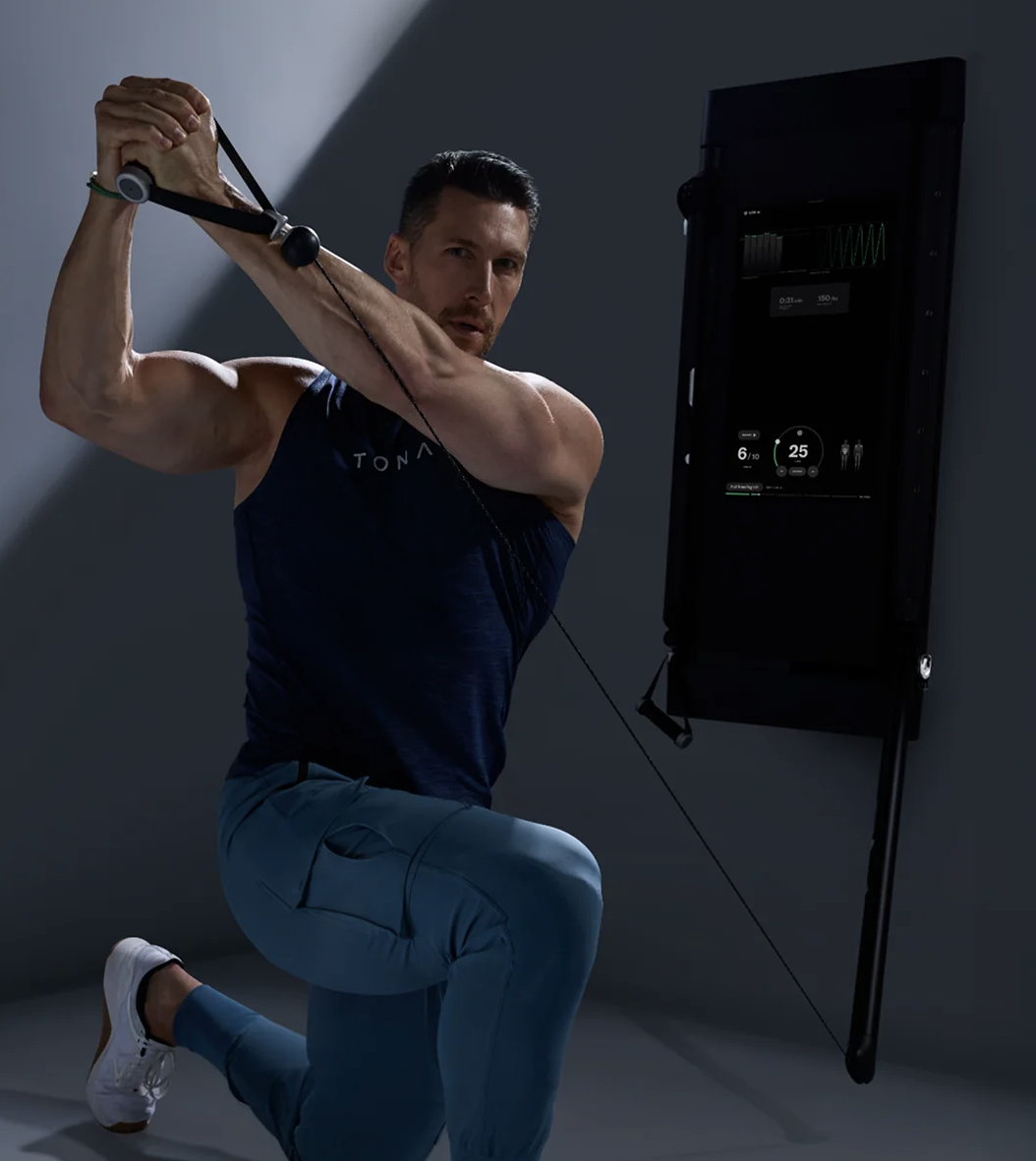 A man performing a resistance exercise using a pulley system with a display monitor showing workout data. He is working out on a home Tonal Fitness machine. 