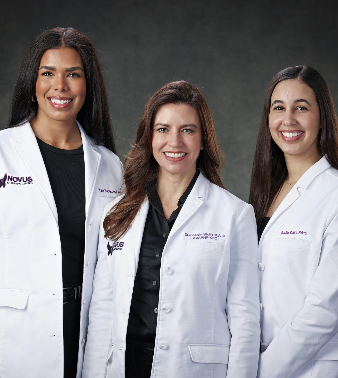 Three women wearing white medical coats with office names and titles, standing together against a dark background, smiling at the camera. They are PAs in an integrative medical center for sexual wellness, HRT, and men's ED services.