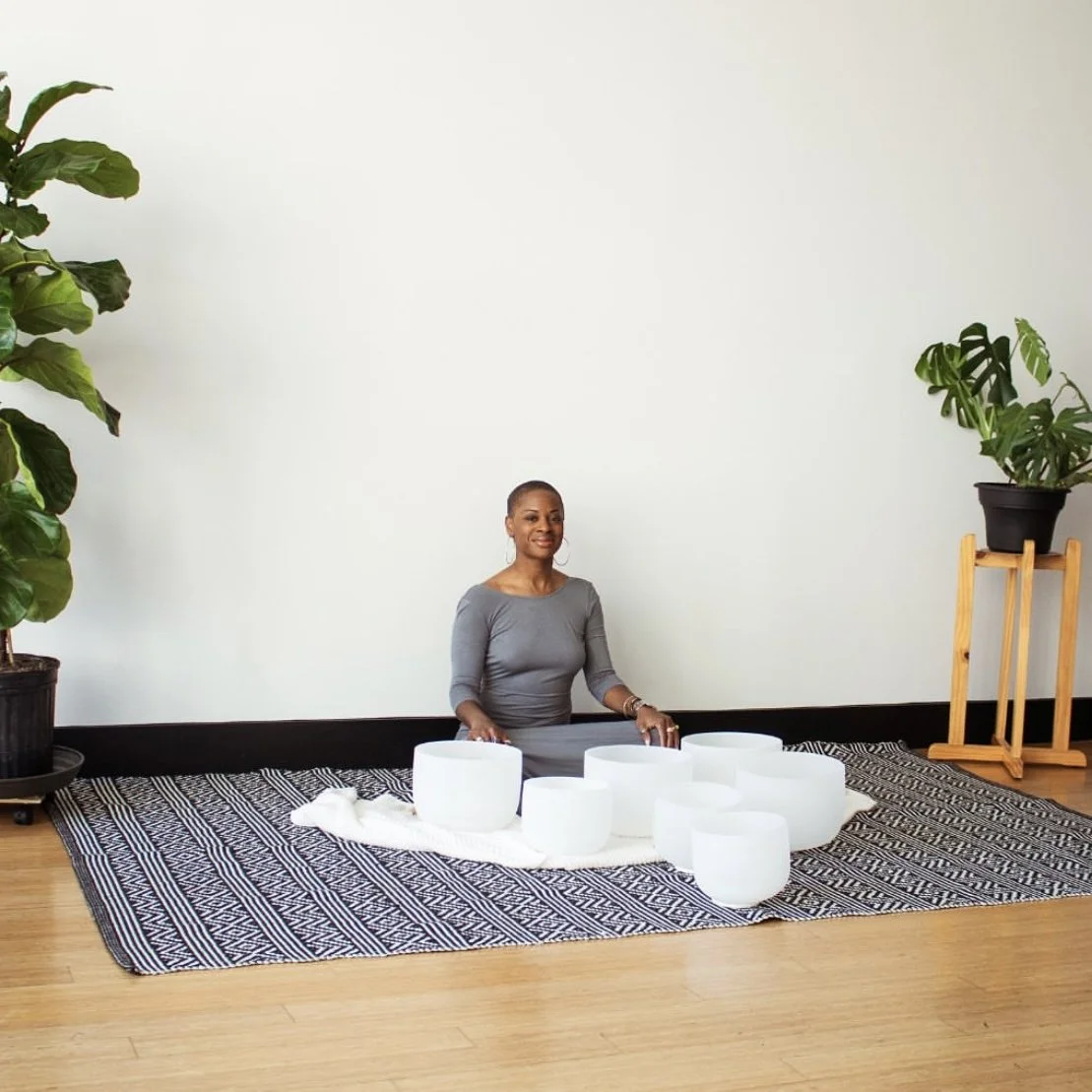 A woman sitting on a patterned rug with white crystal singing bowls in front of her, flanked by potted plants on either side. A meditation coach in Los Angeles 
