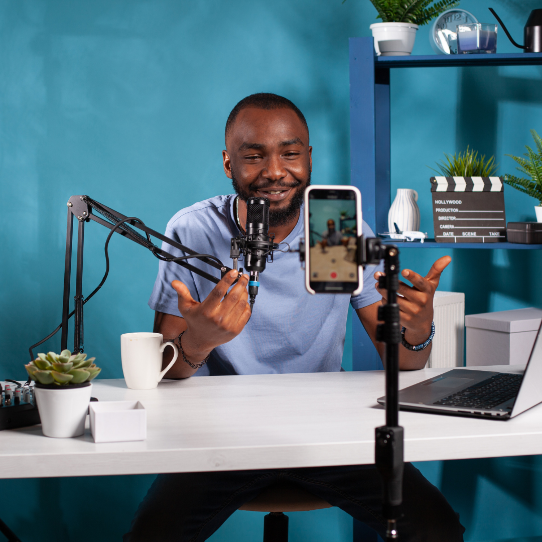 male creator in content creation mode with iPhone and plant on desk and blue background. He is doing a talking head video and working on his personal brand.