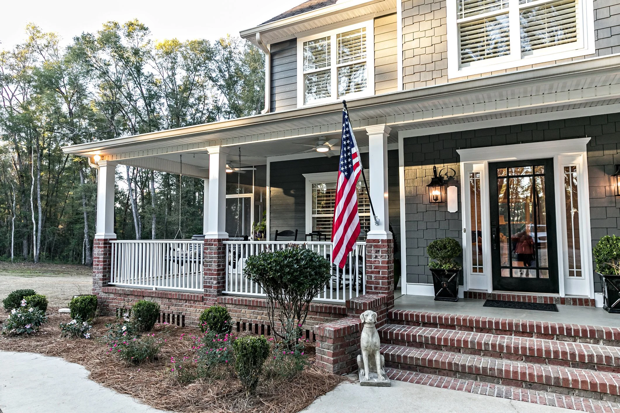 Front porch of a house with a flag, potted plants, and a granite dog statue, surrounded by a garden with bushes and flowers, and trees in the background.