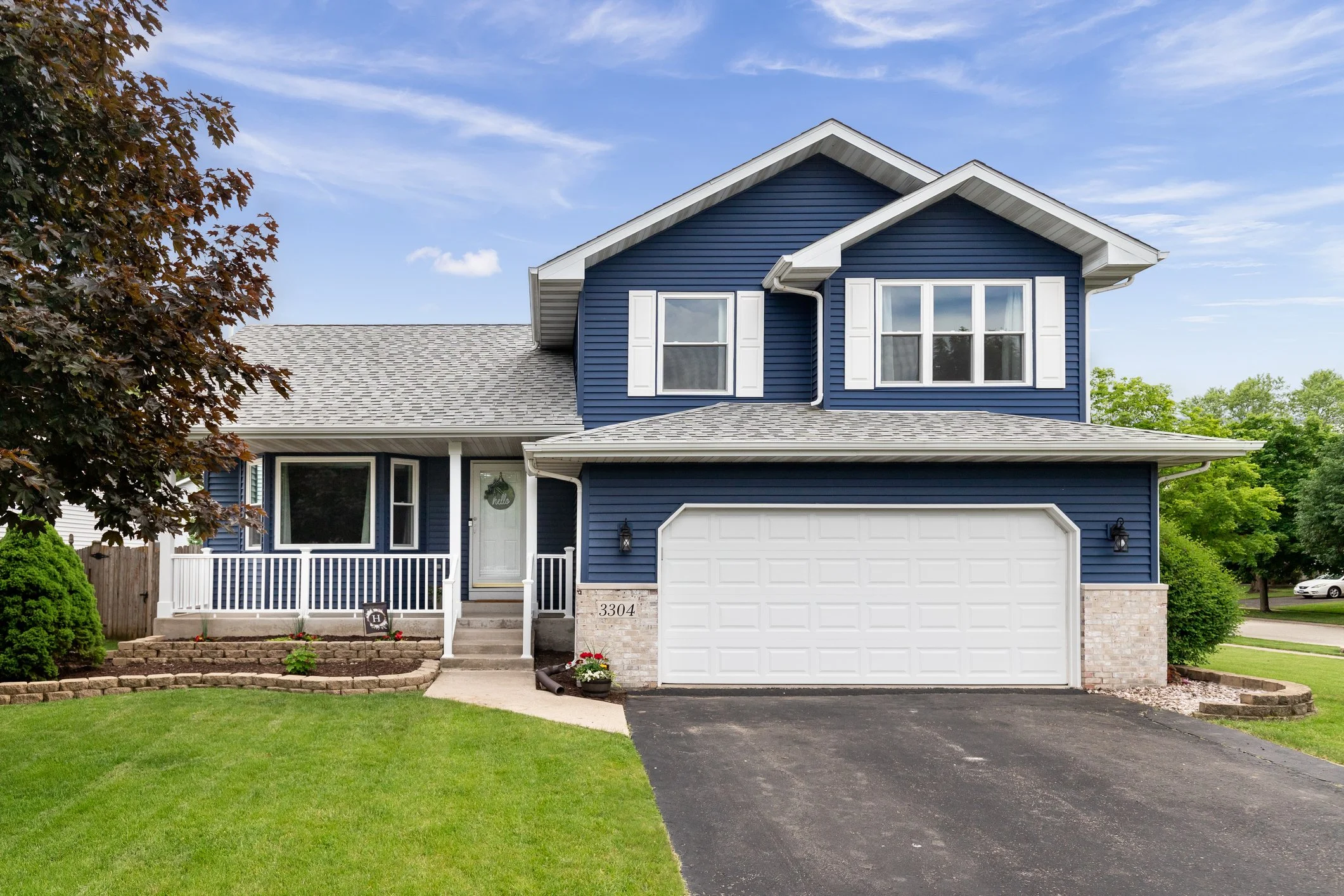 Front view of a blue two-story house with white trim, a garage door, a concrete walkway, a small front porch with white railing, and a lawn with bushes and a tree on the left. The house has a gray shingle roof, and there are other houses and trees visible in the neighborhood. The sky is partly cloudy with some blue sky.