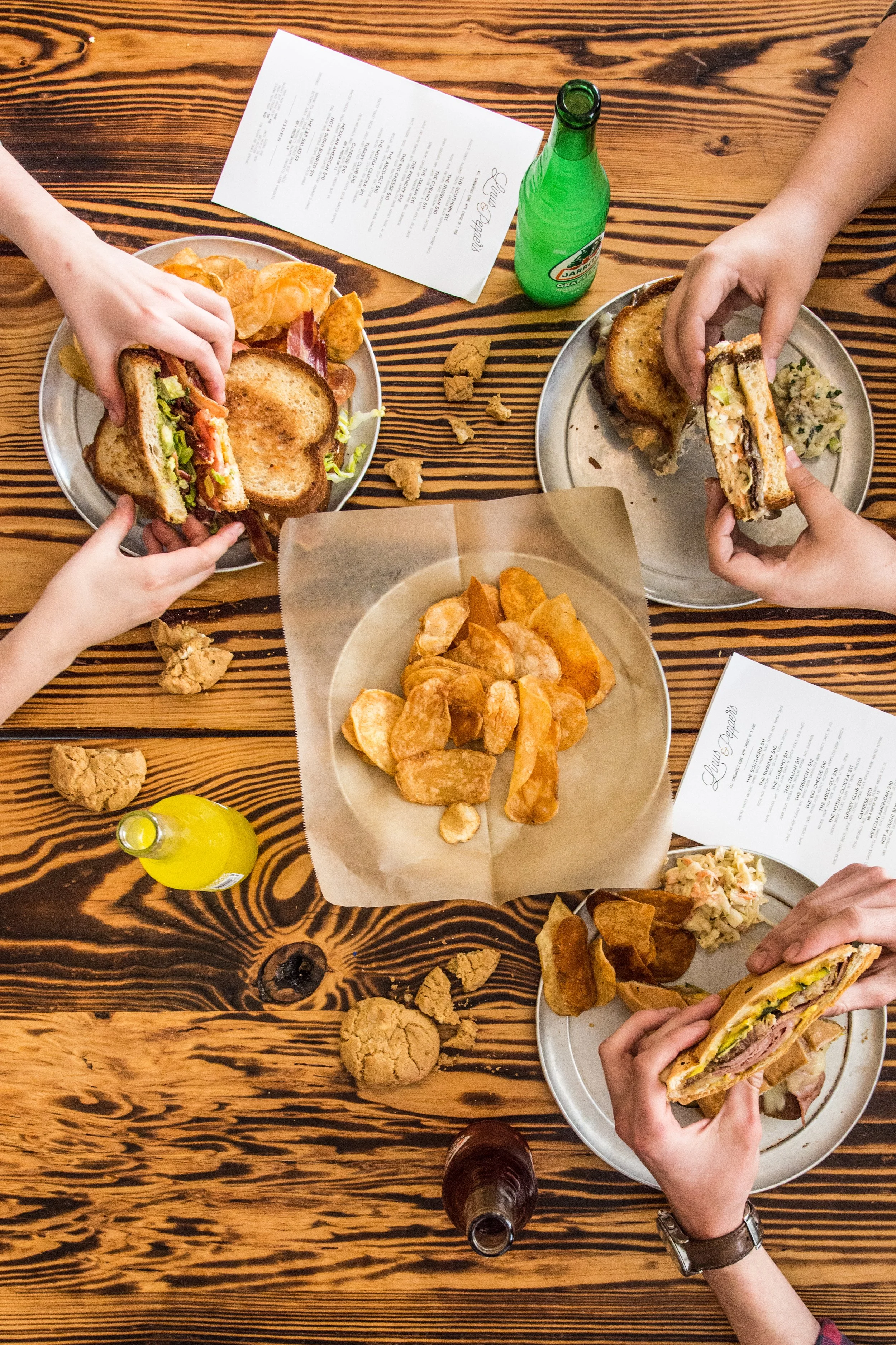 Overhead shot of a wooden dining table with three plates of sandwiches, potato chips, and coleslaw. There are two unopened soda bottles, one green and one brown, and scattered pieces of fried chicken on the table.