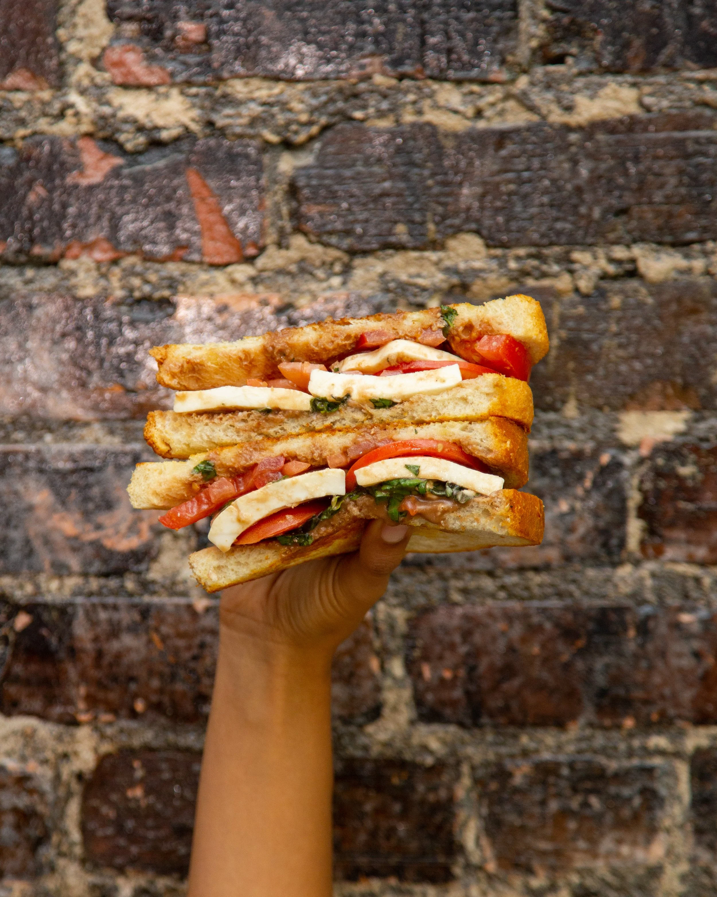 Hand holding a large grilled sandwich with tomatoes, mozzarella, basil, and sauce against a brick wall background.
