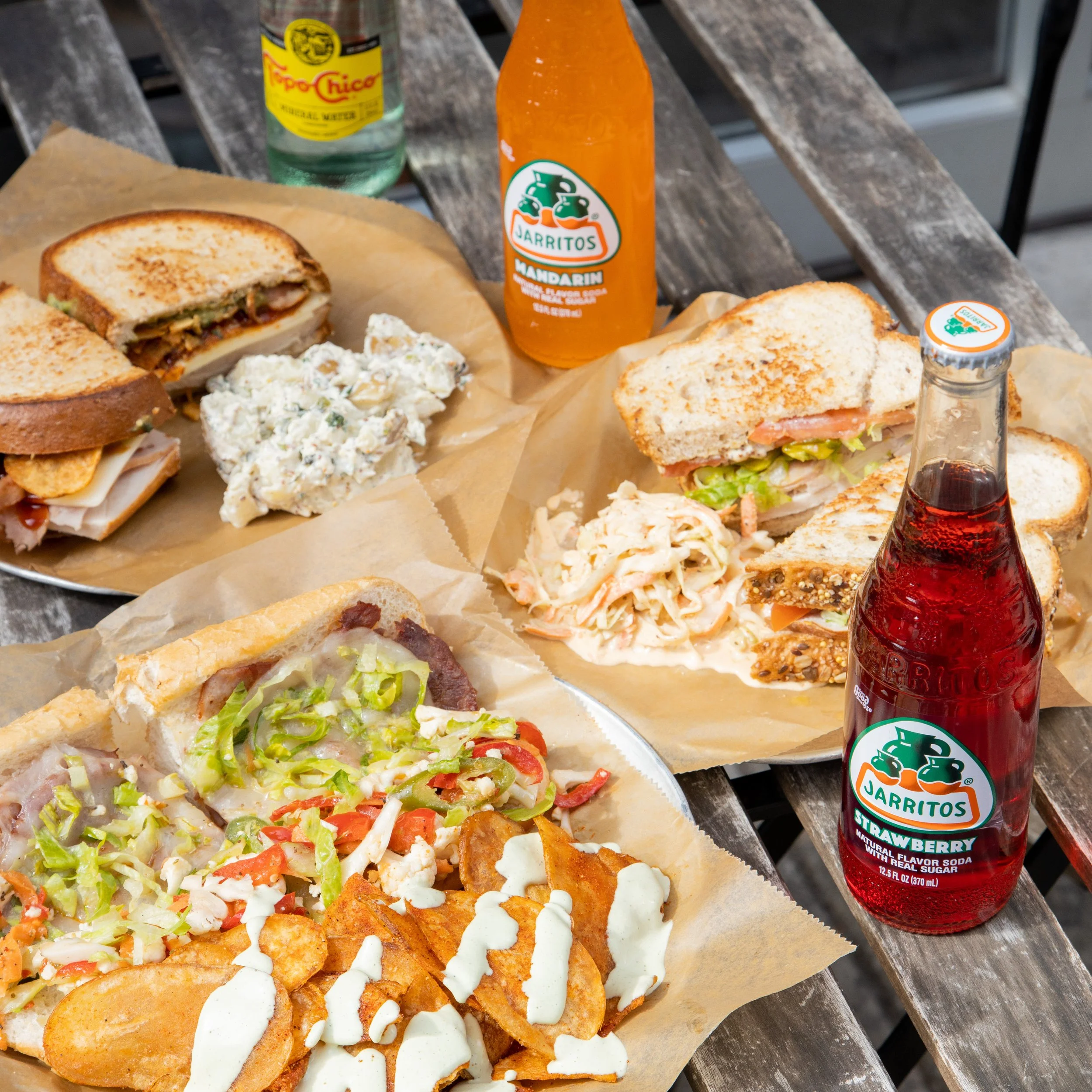 A table with three plates of sandwiches, two jars of Jarritos soda (mandarin and strawberry flavors), a bottle of Topo Chico sparkling water, and a serving of potato chips with white sauce. The sandwiches contain various fillings like meats, lettuce,