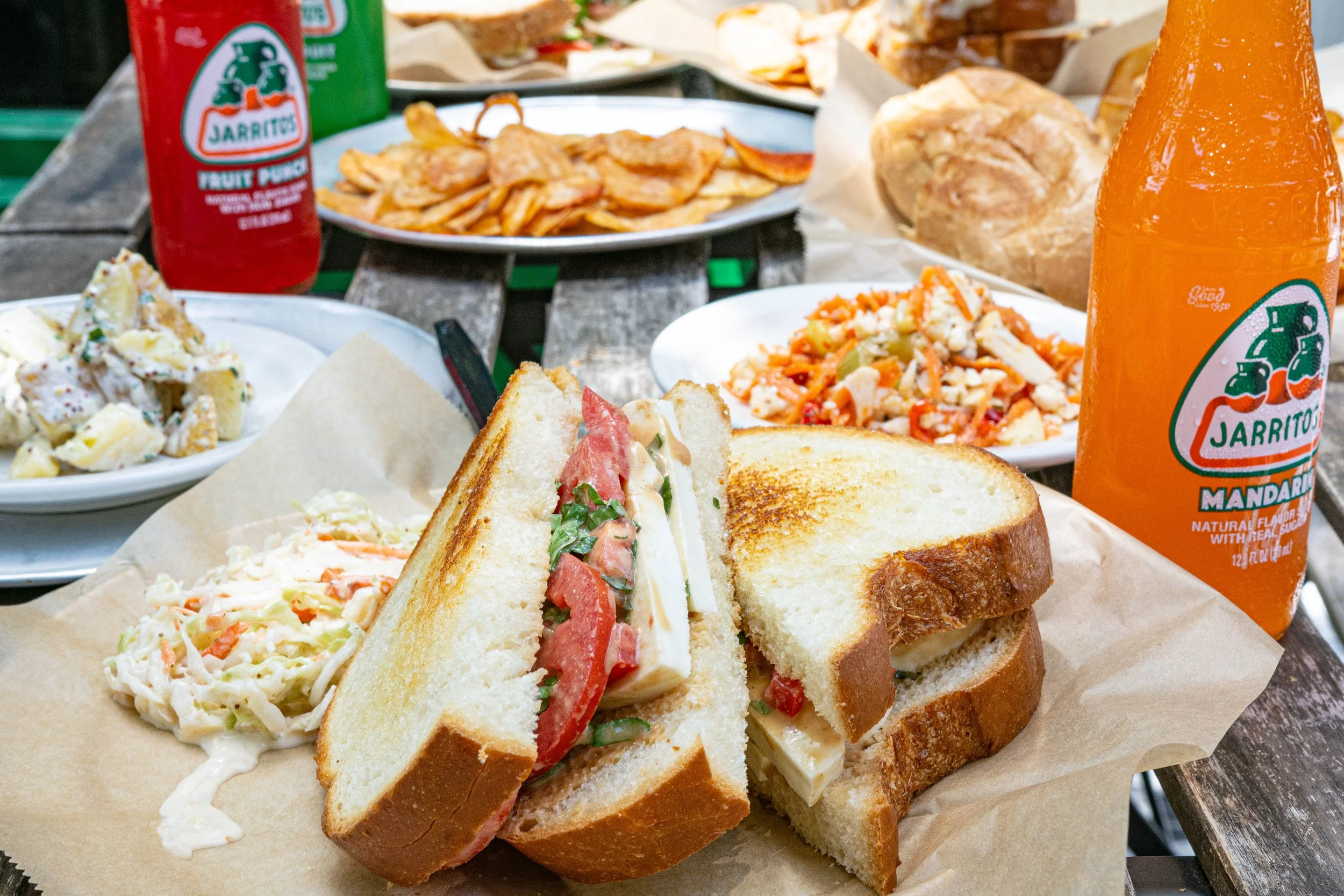 Keto sandwich with tomato, lettuce, and cheese on toasted white bread in a paper-lined basket, surrounded by potato salad, pasta salad, chips, and bottled Jarritos mandarin soda on a rustic outdoor table.
