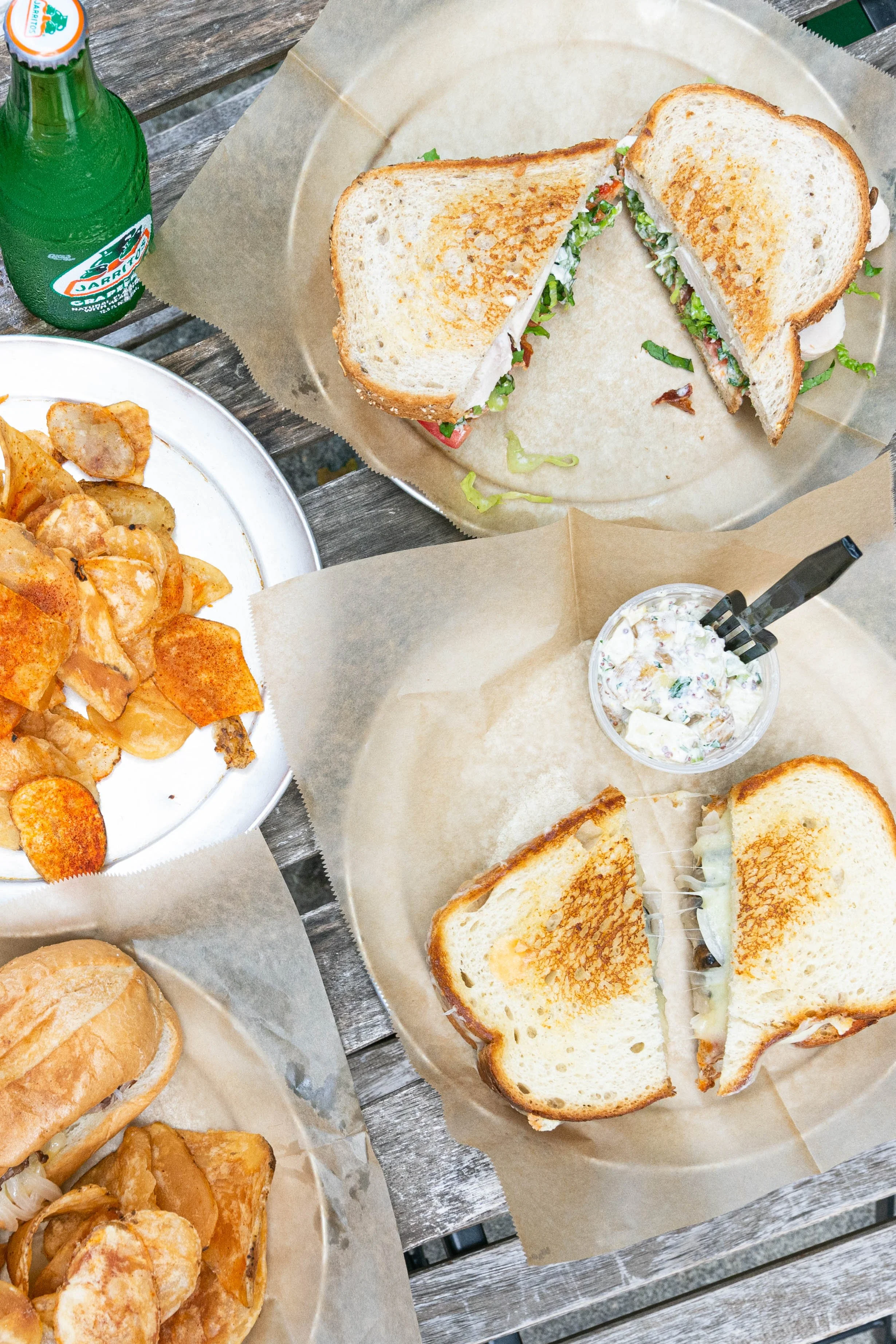 Two halves of a grilled chicken sandwich with lettuce and tomato, a bowl of potato chips, a sandwich with melted cheese and onions, and a bottle of Sprite on a wooden table.