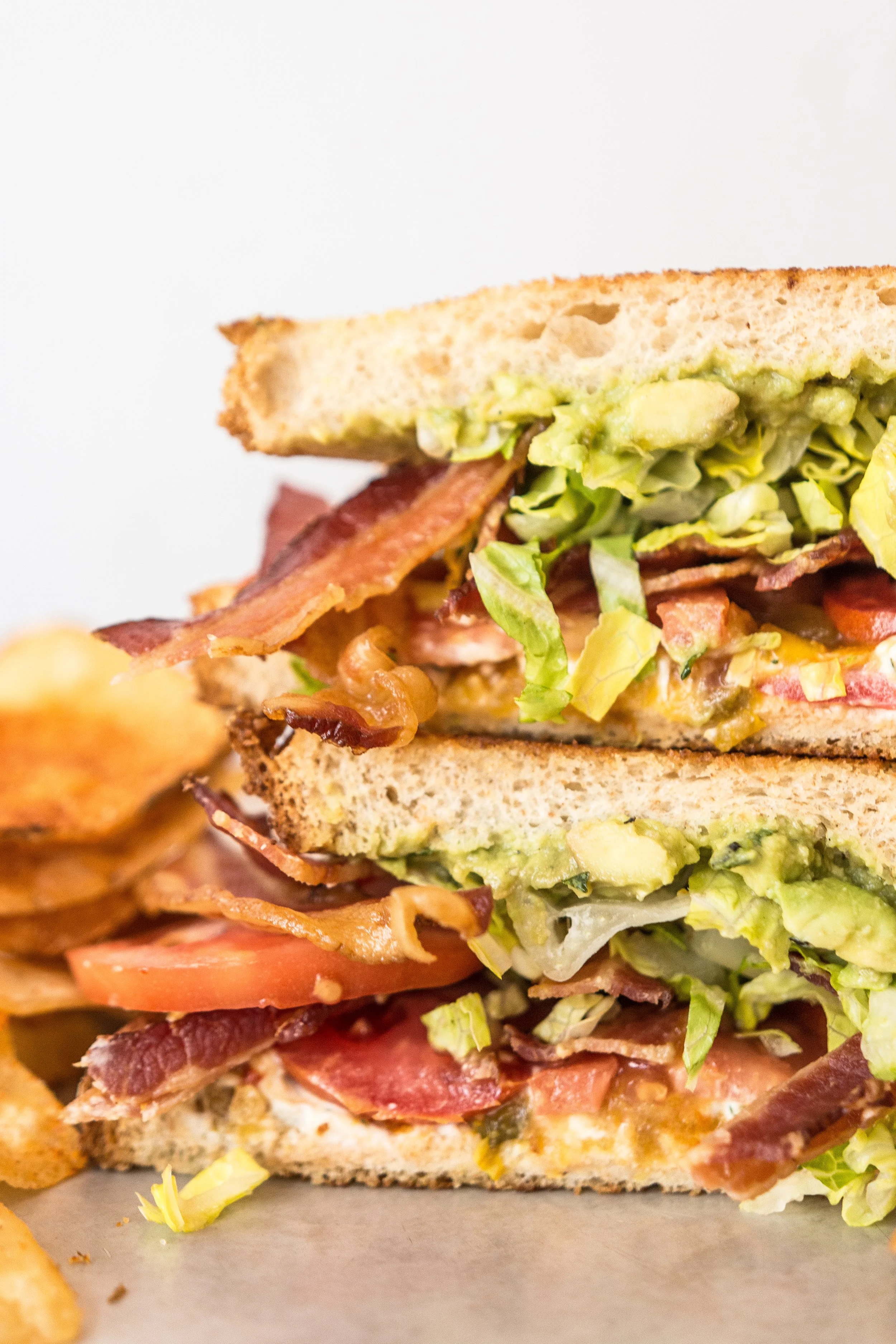 Close-up of a double BLT sandwich with bacon, lettuce, tomato, and avocado on toasted bread, accompanied by potato chips.