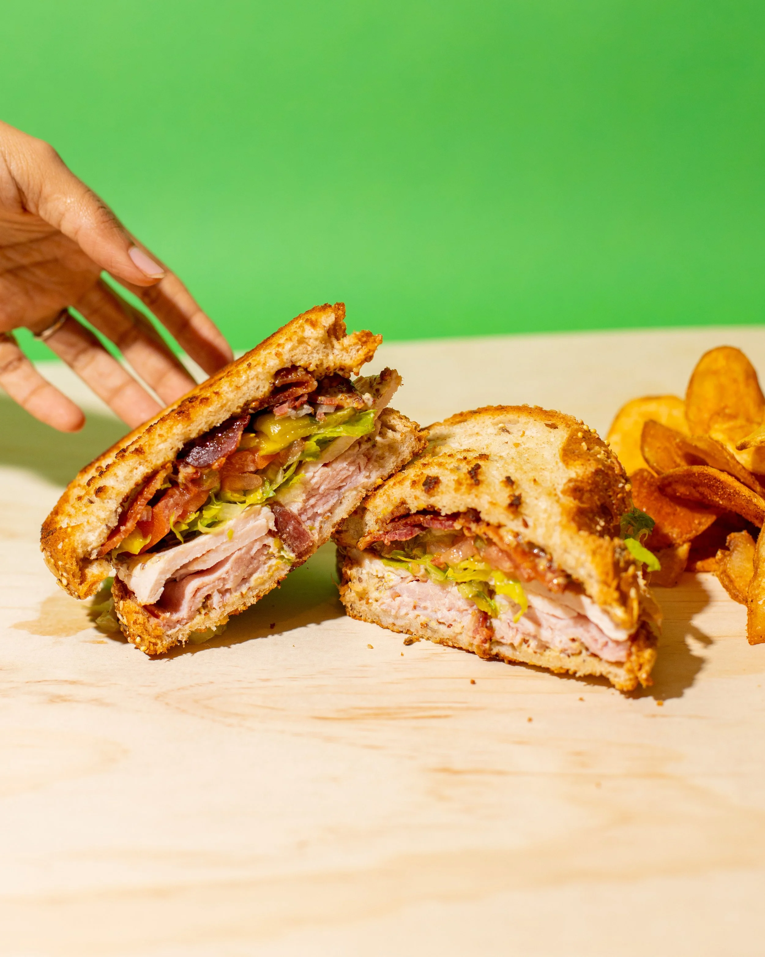 Halved fried chicken sandwich with lettuce, tomato, and bacon, served with potato chips on a wooden surface against a green background.
