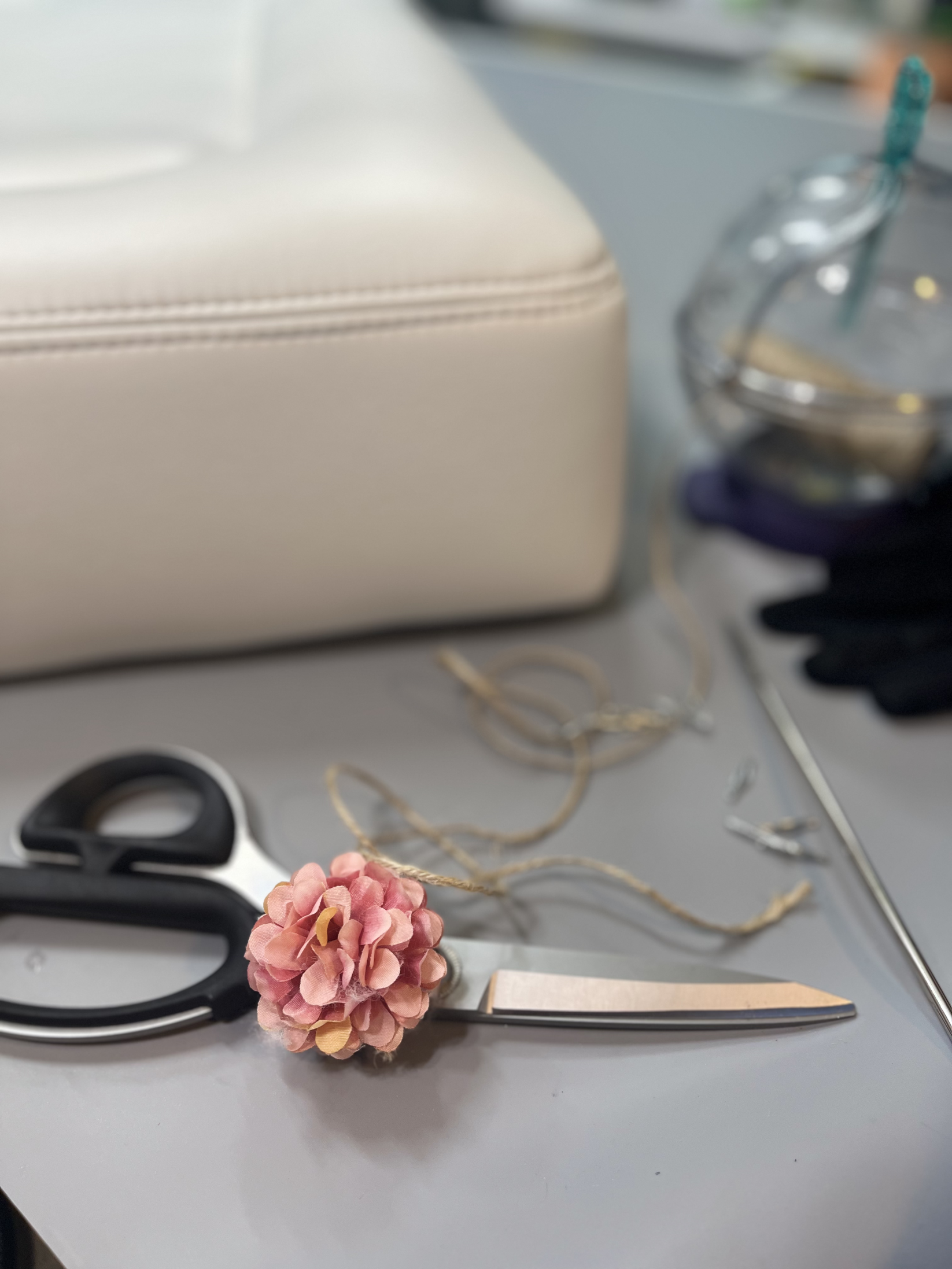 Scissors with a pink decorative flower on the handle, on a gray surface with various objects including a beige bag, a glass with a straw, and gloves.