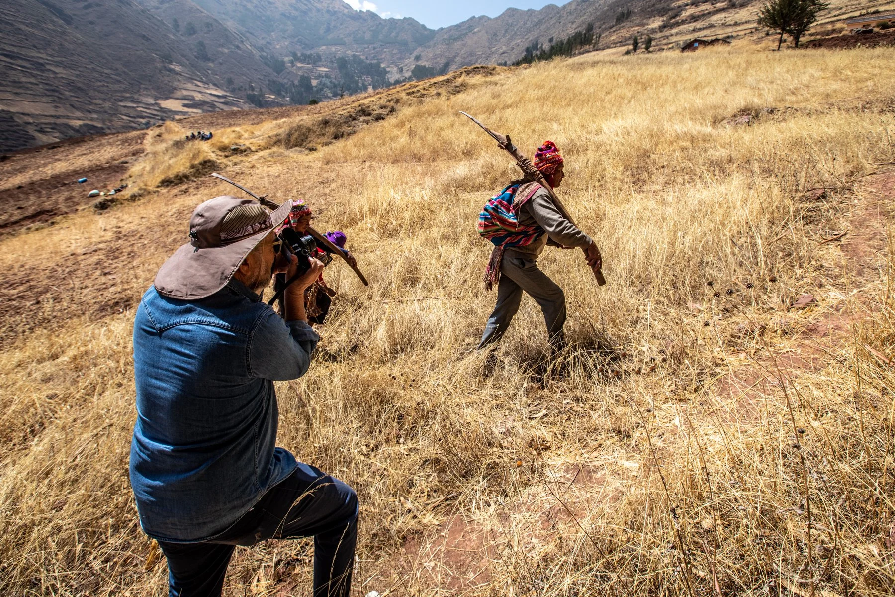 Un grupo de personas en un paisaje montañoso, con dos personas en primer plano disfrazadas con atuendos tradicionales indígenas, mientras que otra grabadora captura la escena, en un entorno de hierba seca y colinas en el fondo.