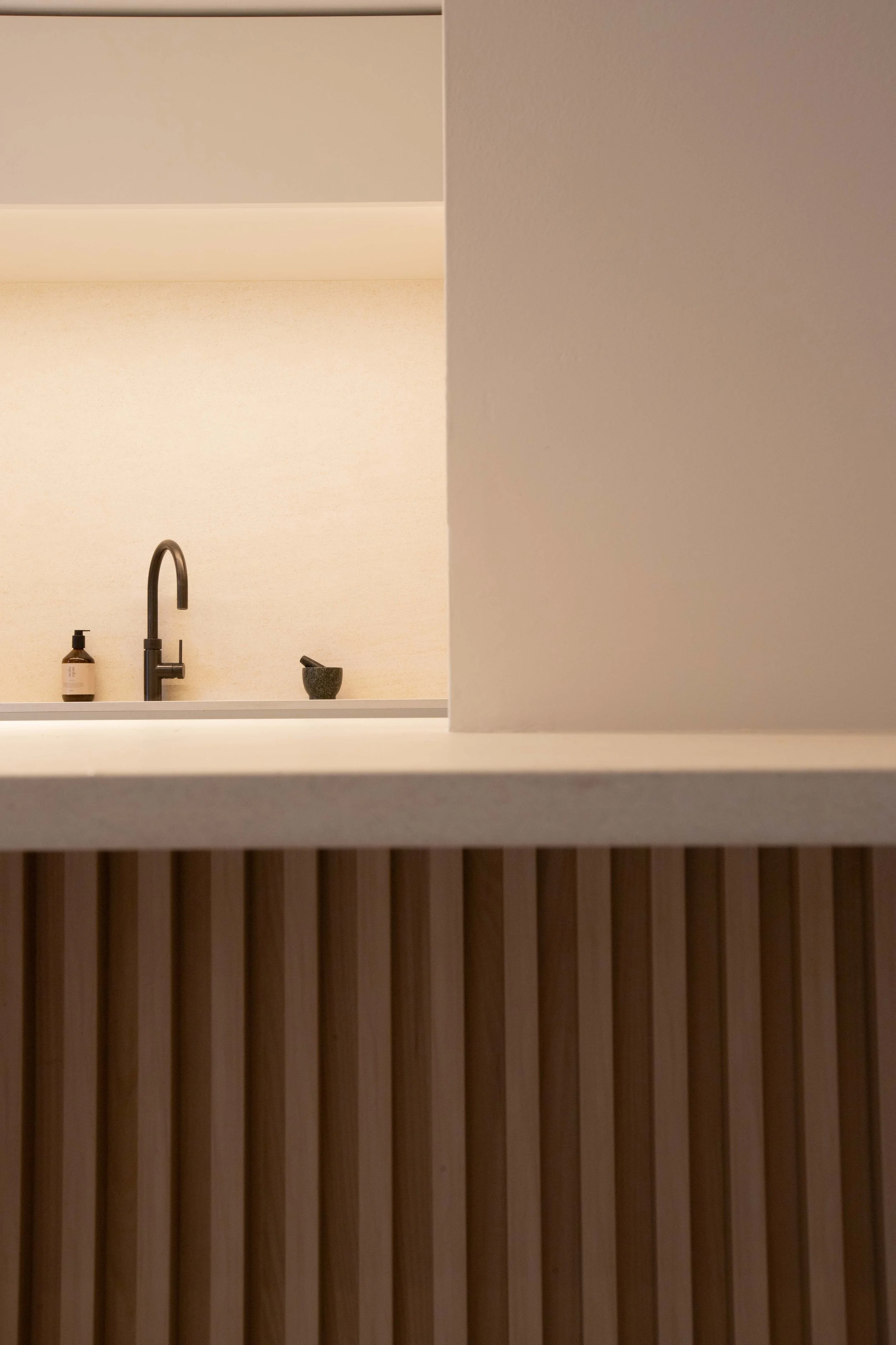 Close-up of a minimalist kitchen or bathroom counter with a black faucet, a soap dispenser, and a small bowl on a beige wall in the background, featuring vertical wooden slats in the foreground.