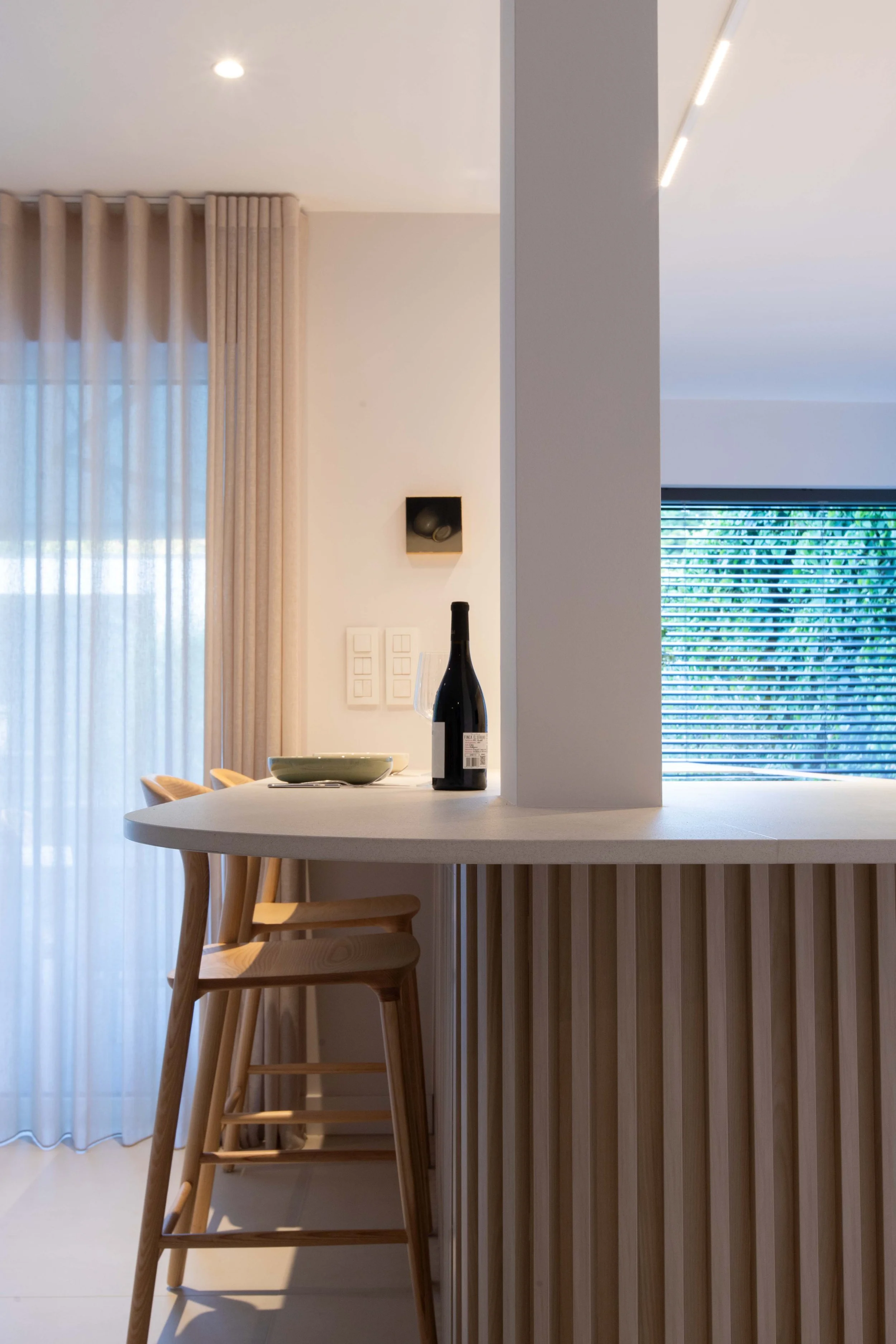 Modern dining area with a wooden bar-height chair, a bottle of wine, a steering glass, a bowl, and a partial view of a window with blinds and curtains.