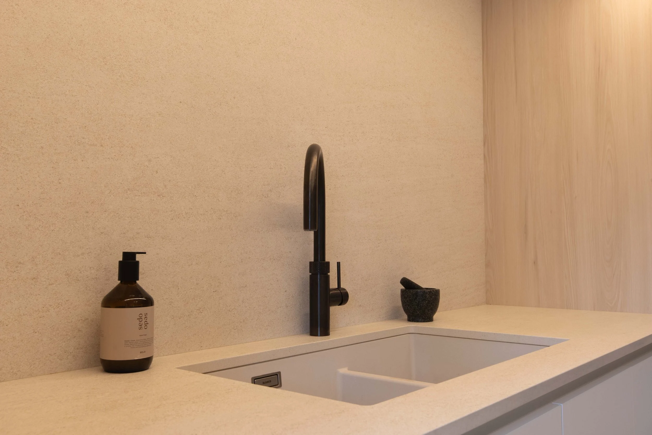 A modern kitchen sink area with a black faucet, a bottle of soap, a mortar and pestle, and a beige countertop with a textured beige wall background.