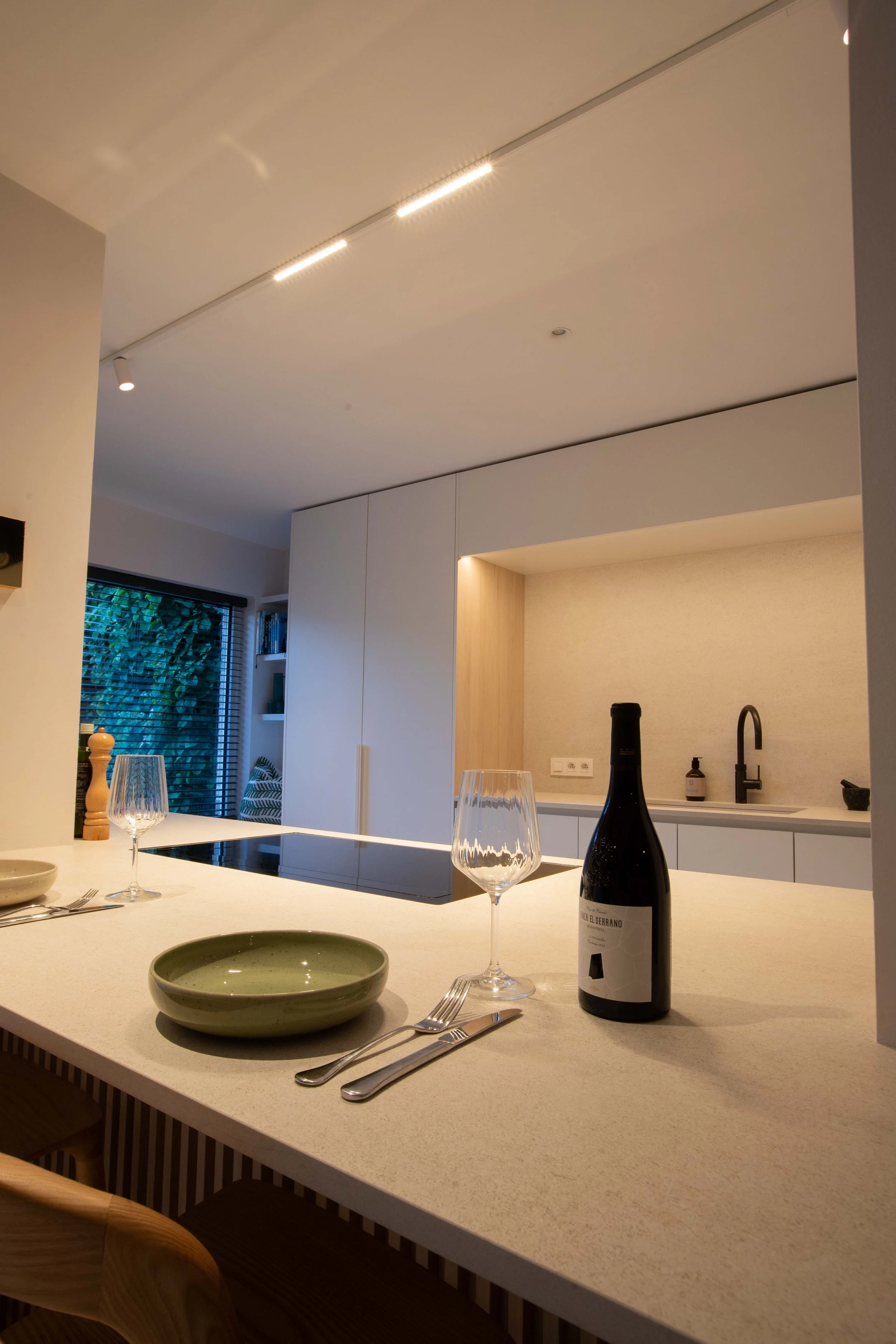 A kitchen with a white countertop set for a meal, including two wine glasses, a green bowl, and a bottle of wine. In the background, there are white cabinets, a sink, and a window with blinds.
