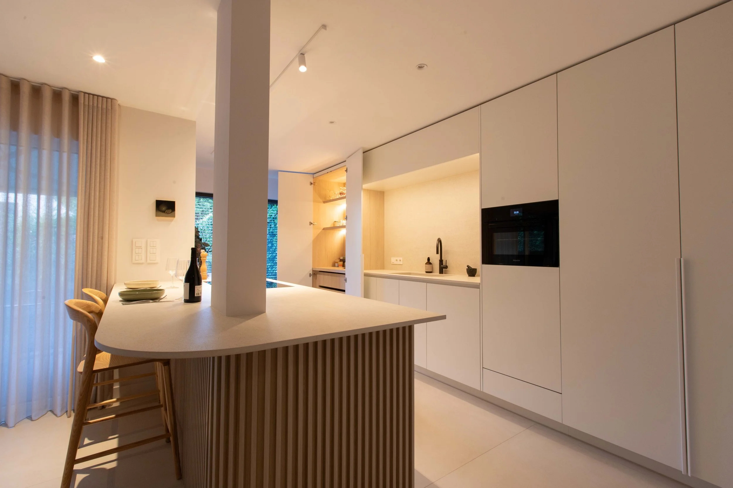 Modern kitchen with white cabinetry, a built-in oven, a black faucet, an island with a curved edge, beige drapes, and a dining area with wine and plates.