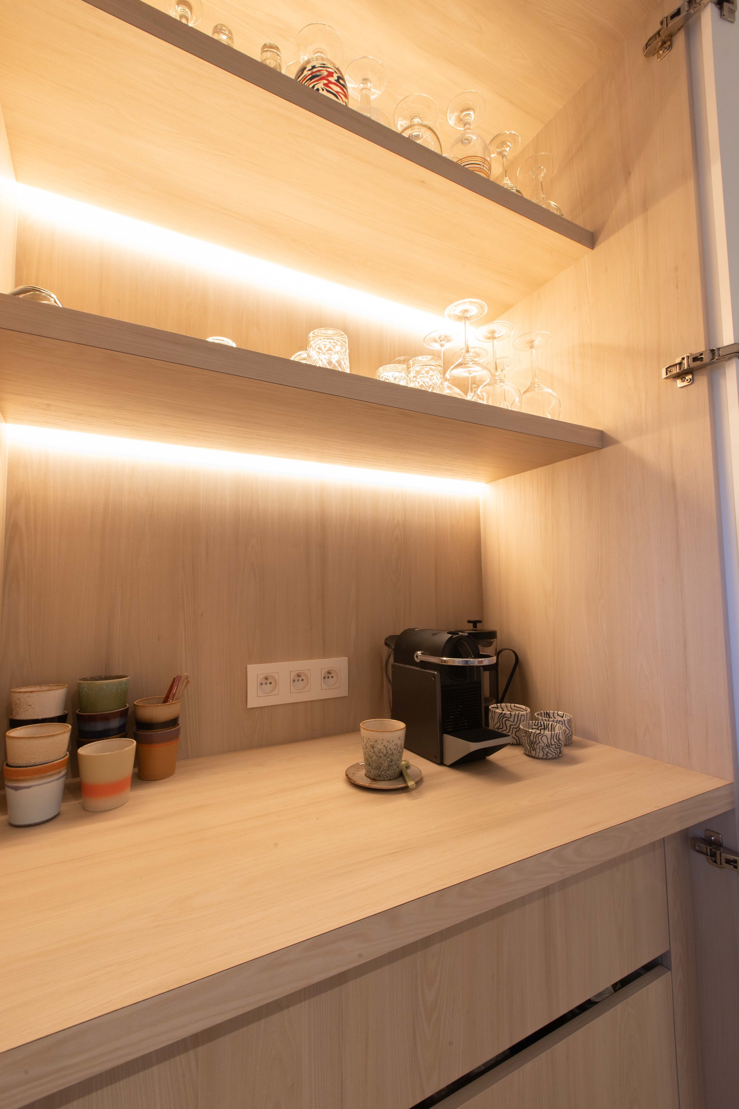 Coffee station with a built-in coffee machine, cups, and glasses on wooden shelves and counter, with LED lighting