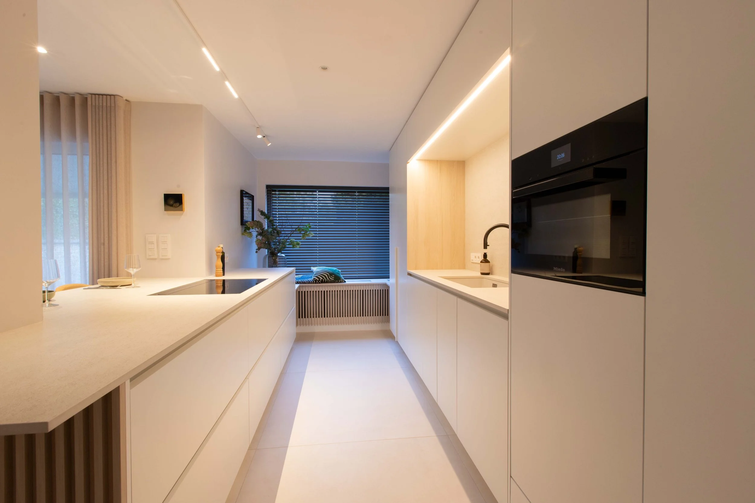 Modern minimalist kitchen with white cabinetry, a built-in oven, a sink, and a window with blinds, decorated with plants and wine glasses.