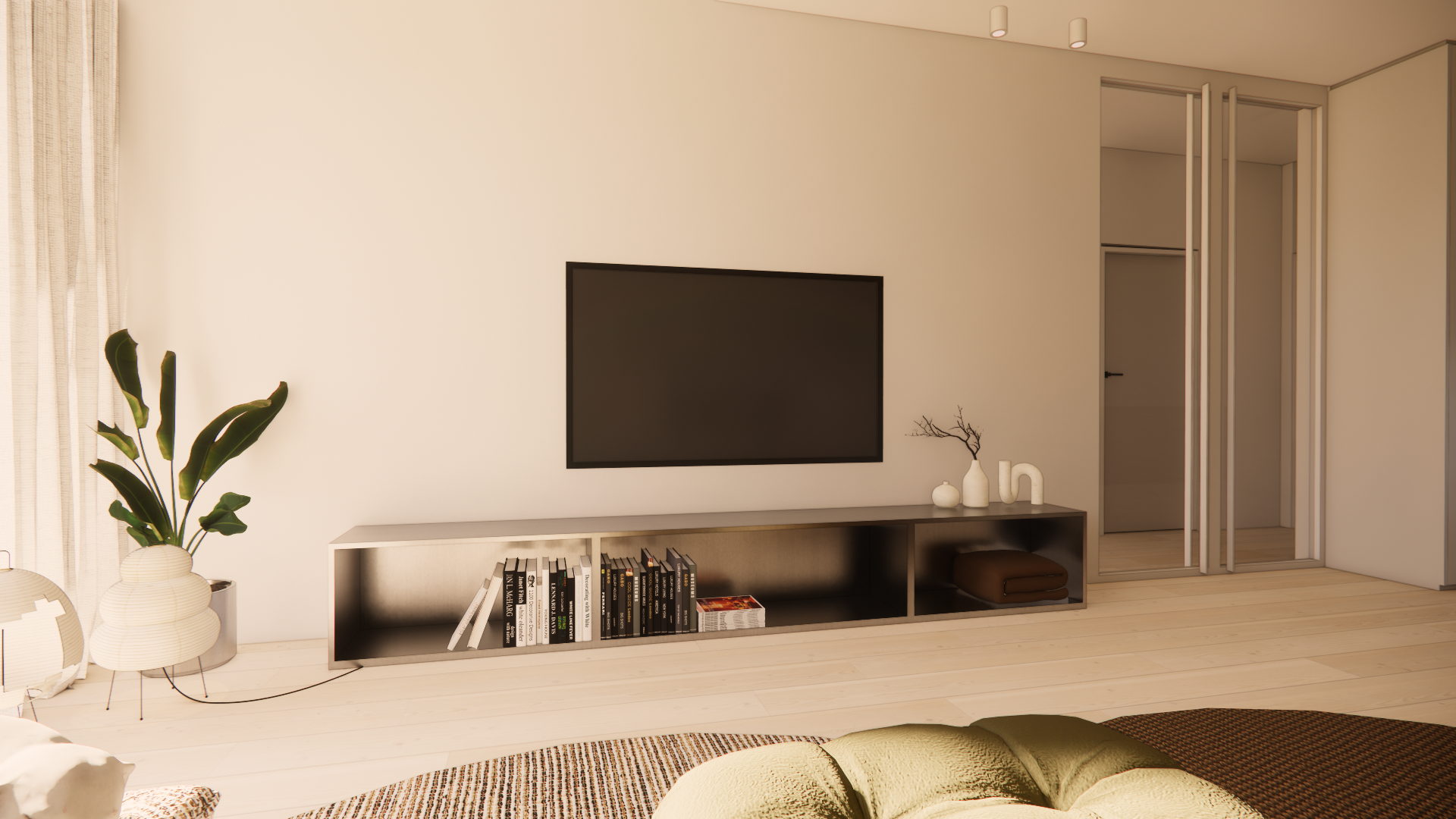 Modern living room with a wall-mounted TV, low white shelving unit with books and decorative items, a large potted plant, and a seating area with cushions. Light-colored wood flooring and neutral walls.