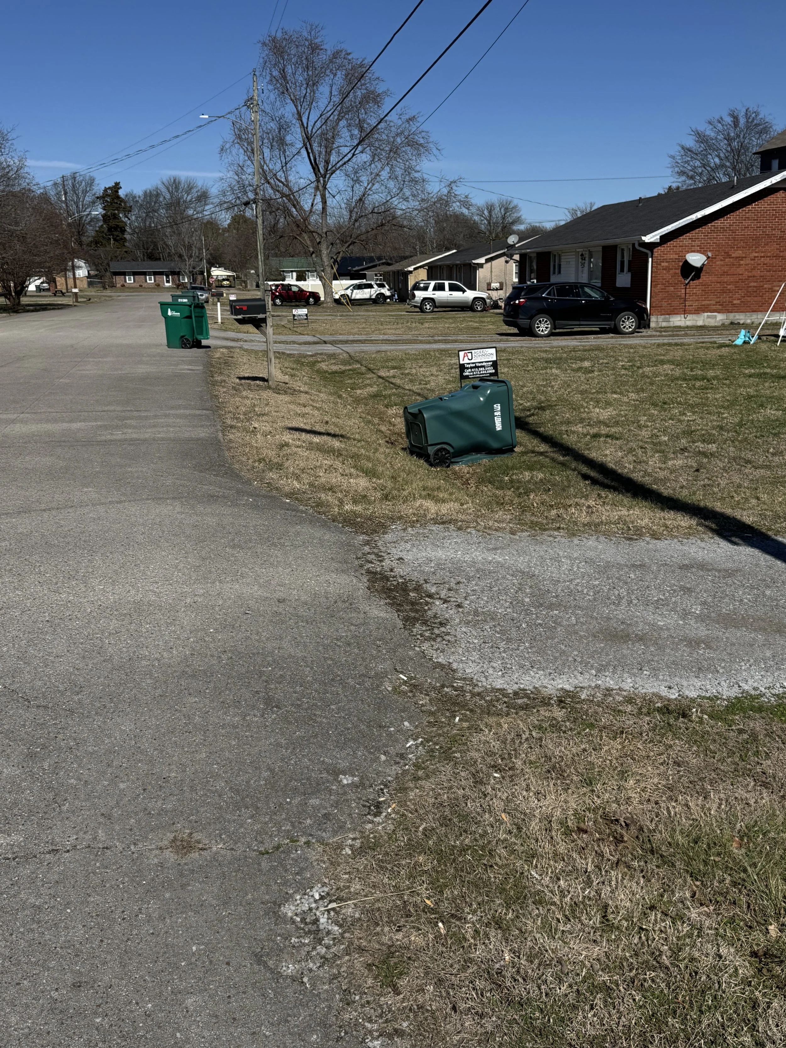 A suburban street with a few houses and parked cars, with two green trash bins in the front yard, and an overturned green recycling bin on the grass.