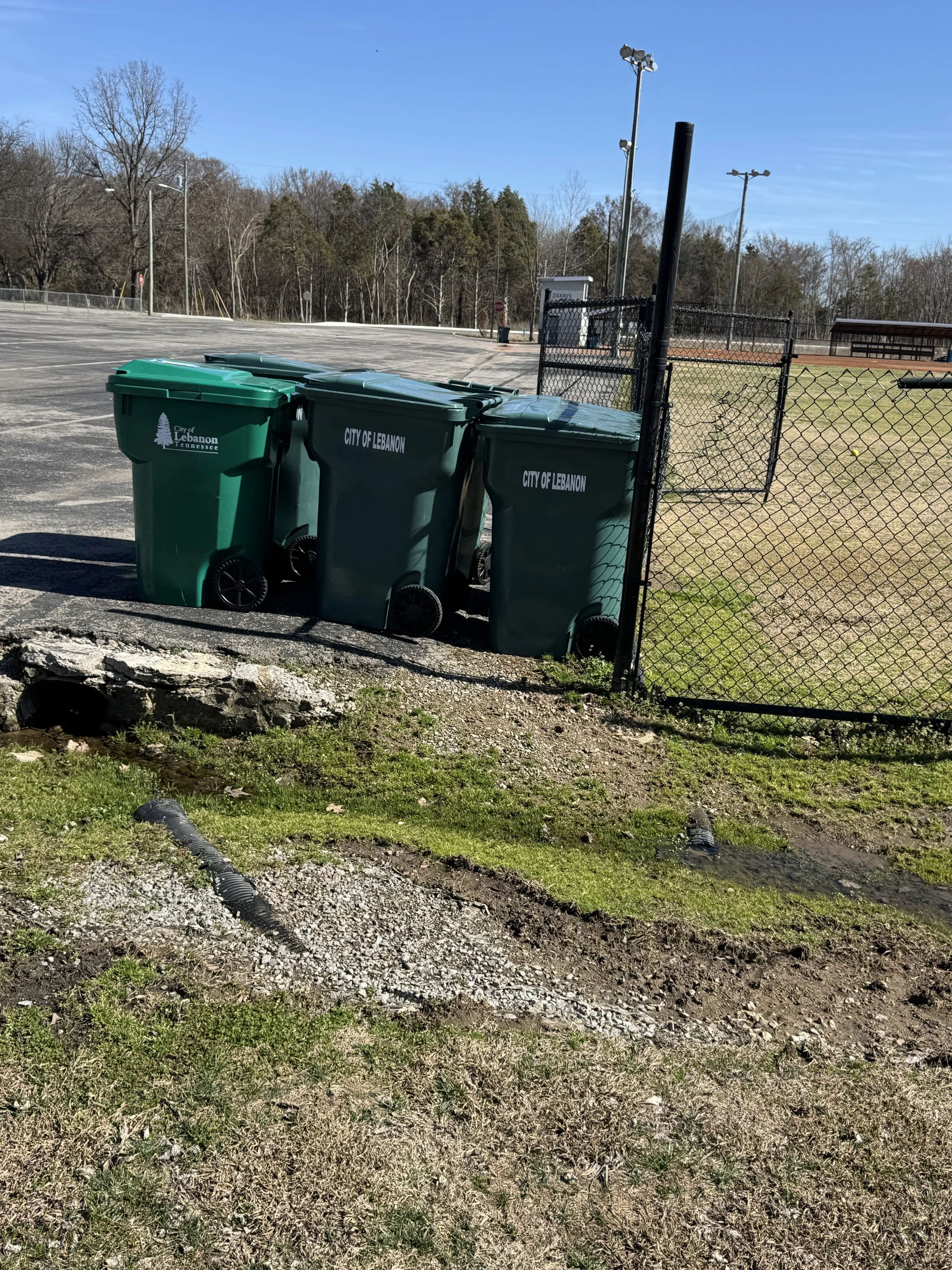Four green trash bins labeled 'City of Lebanon' and 'City of Lebanon Tennessee' outside a gated area, with a baseball field and light poles in the background on a sunny day.