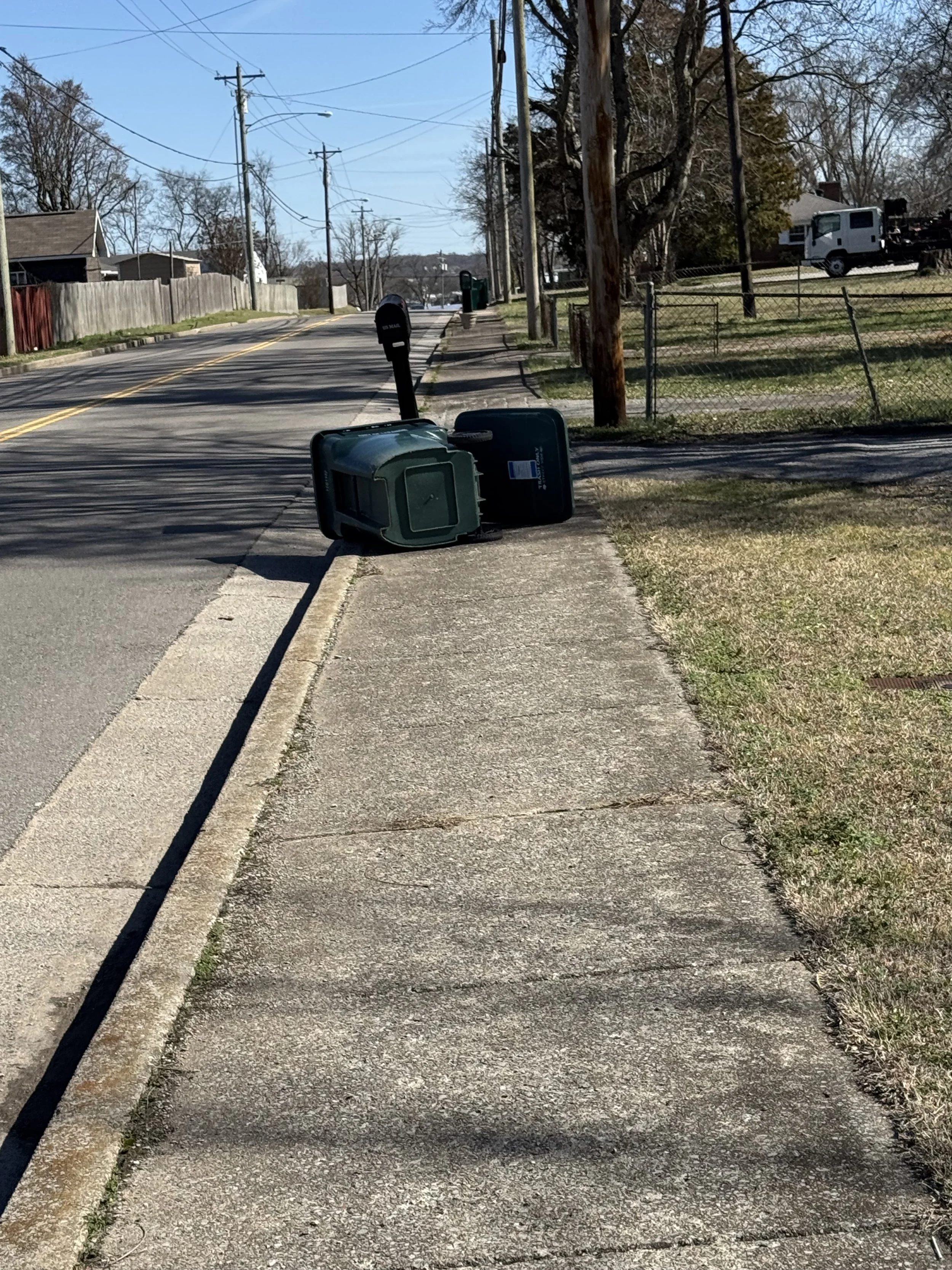 Upside-down trash cans on the sidewalk near the curb, with a mailbox behind them, on a residential street with trees, fences, and utility poles, under a clear blue sky.