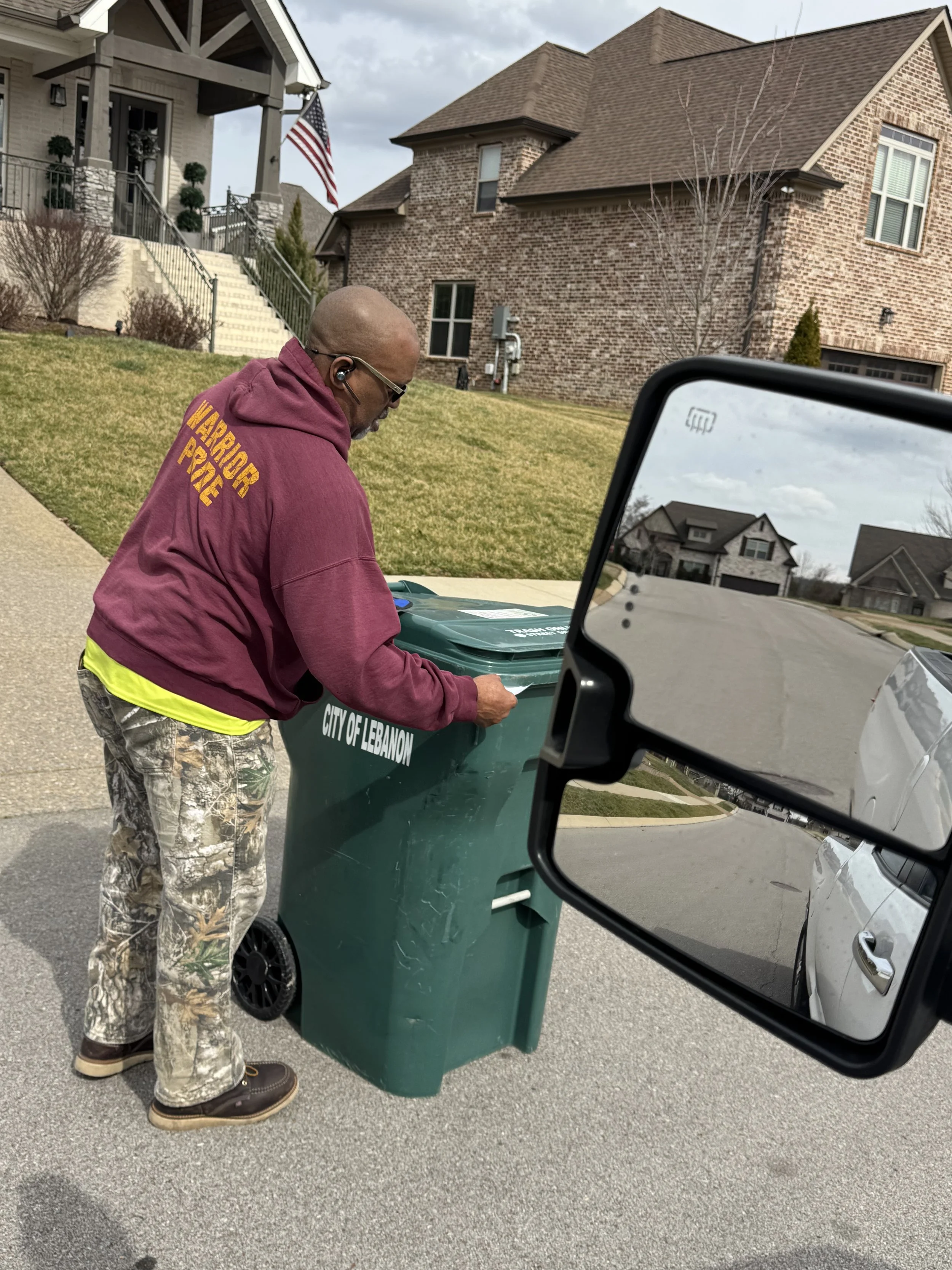 A man in camouflage pants, a maroon hoodie with yellow text, and brown boots is tossing papers into a green city trash can labeled 'City of Lebanon' on a residential street. The photo is taken near someone's side mirror, reflecting houses and a cloud