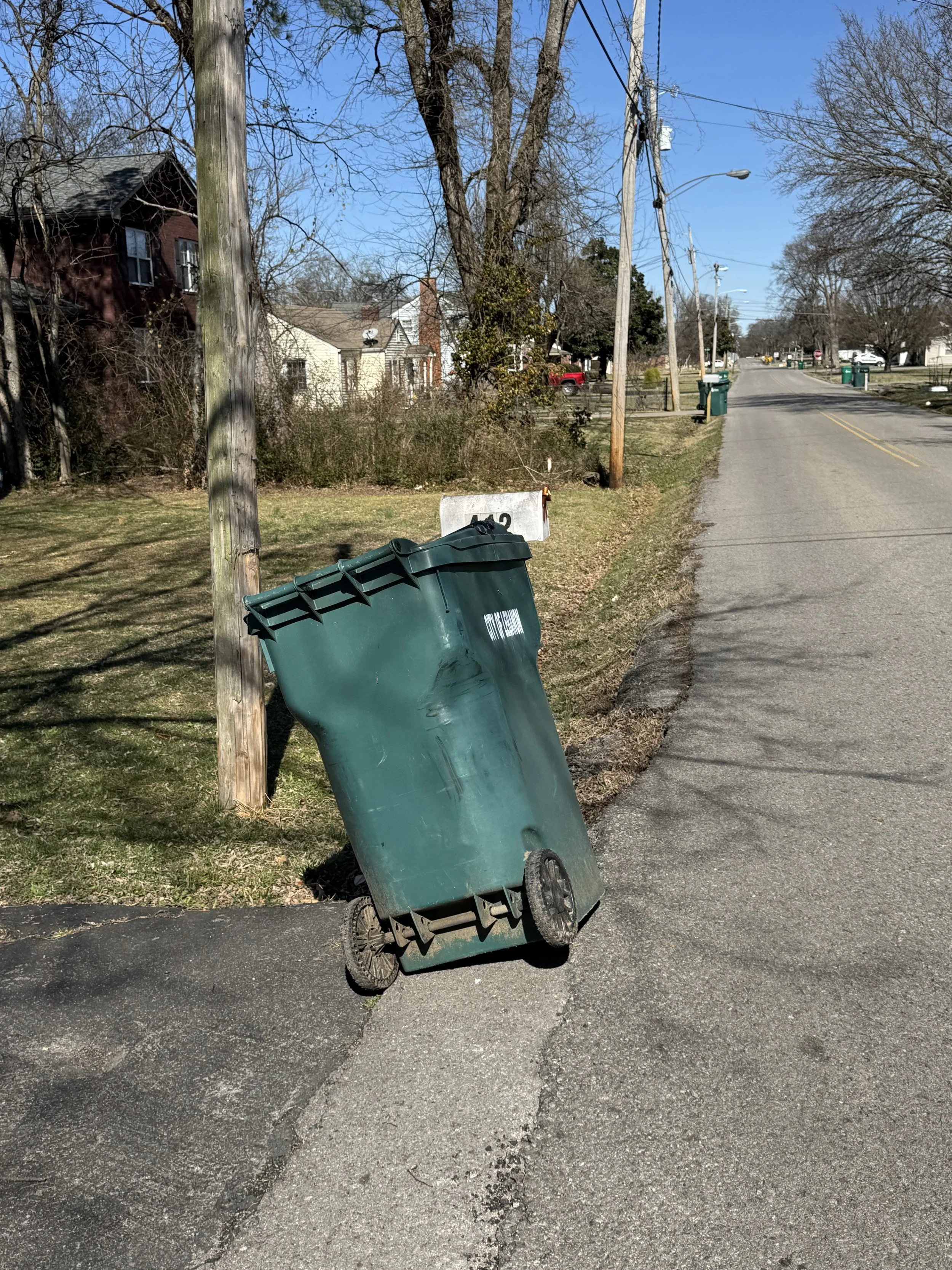 A green trash can is tipped over on the edge of the sidewalk, leaning against a wooden utility pole in a residential neighborhood with houses, trees, and a clear blue sky.