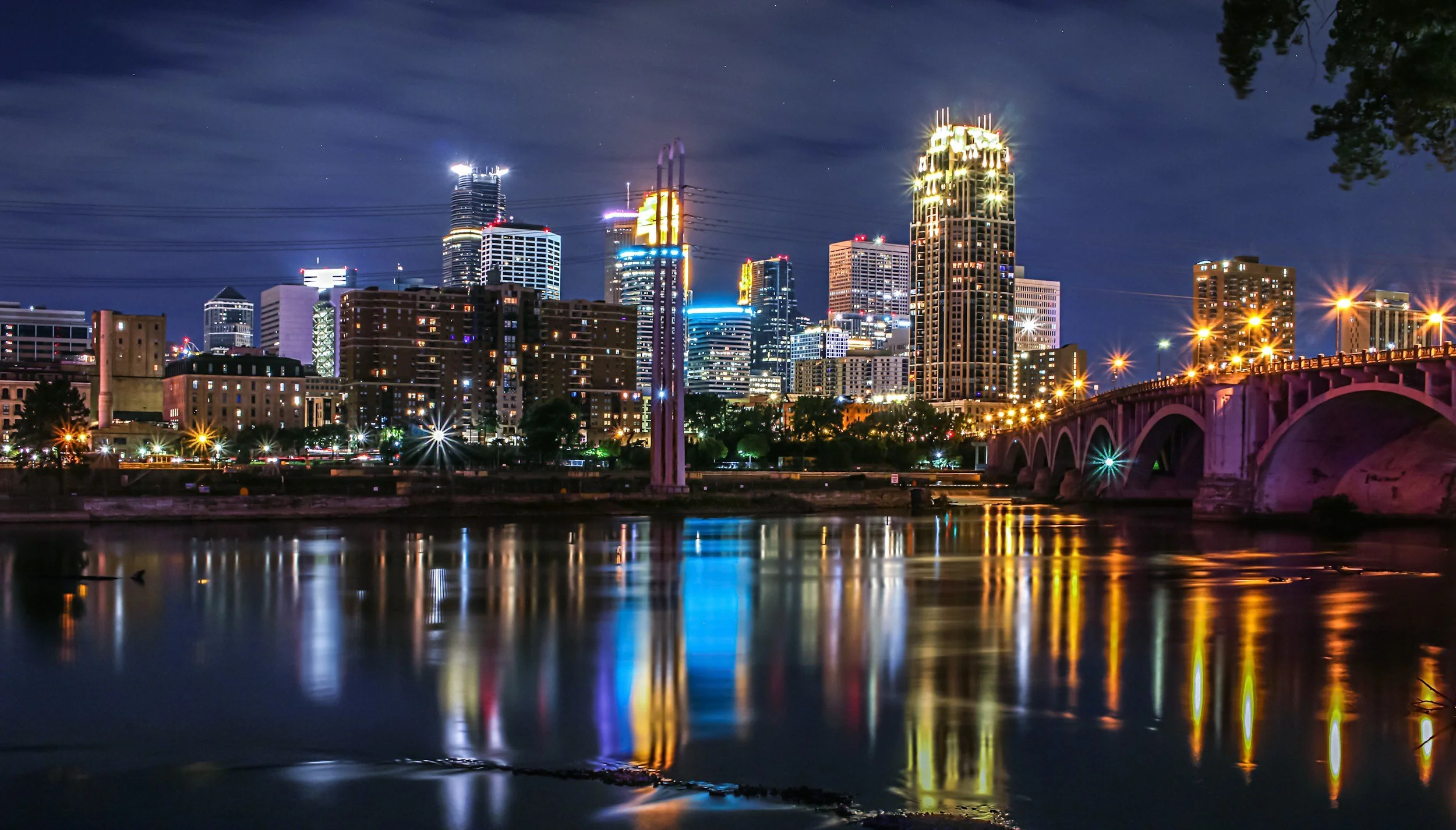 Image of Minneapolis skyline at night. The buildings have bright lights against the sky. The Minnesota River is in the forground.