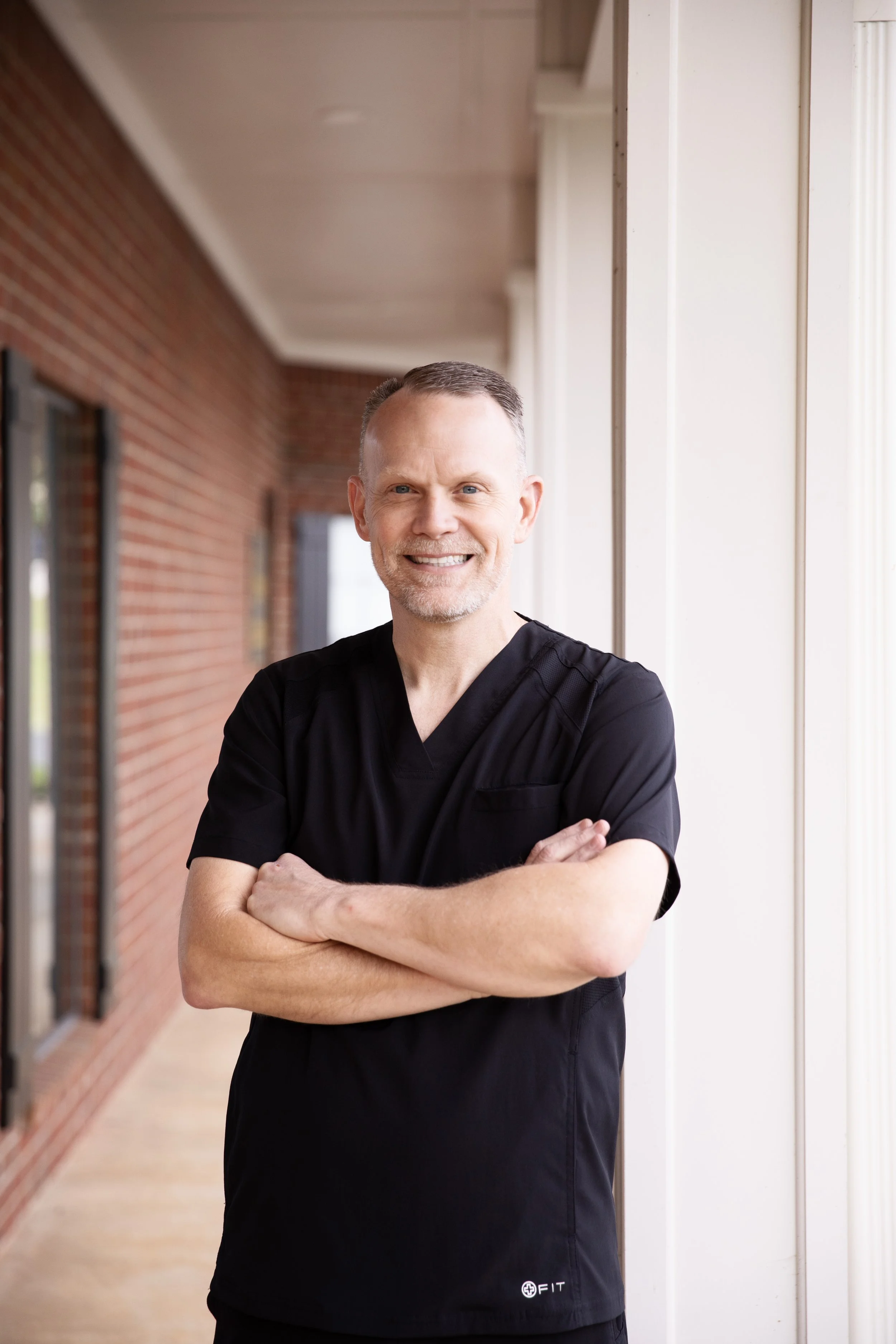 Dr. Hamons smiling at the camera. He is in black medical scrubs and standing outdoors with arms crossed in front of a brick building and large glass windows.