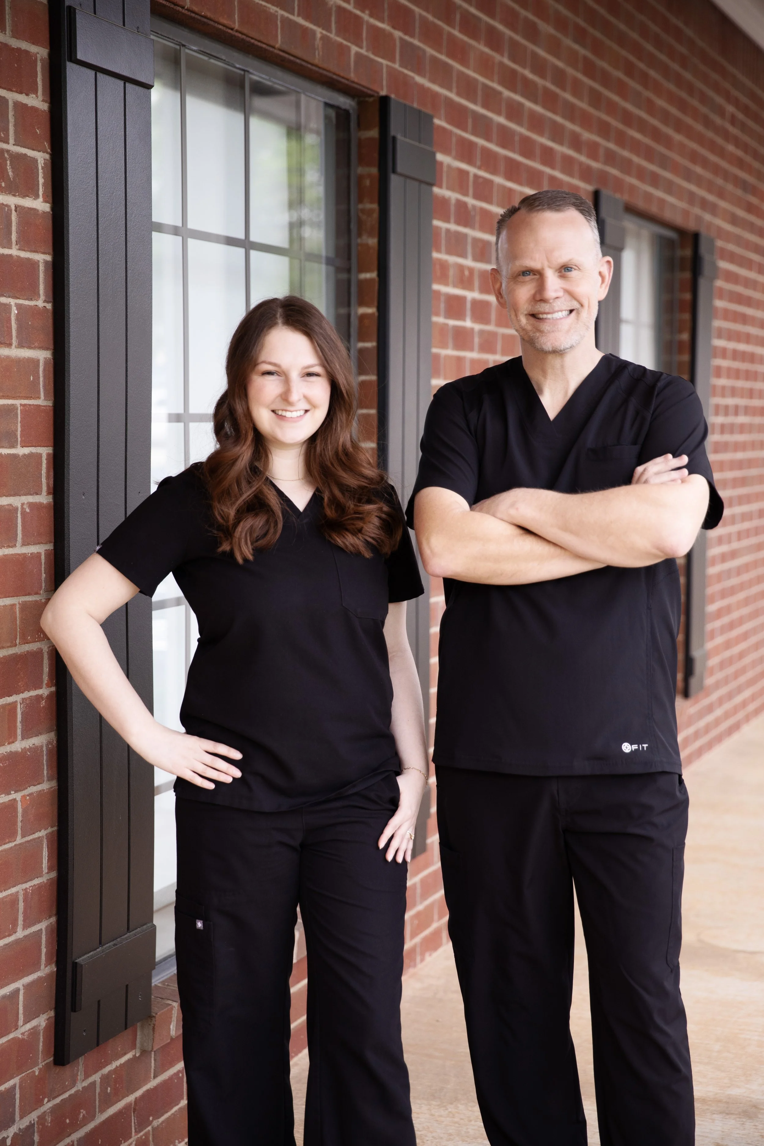 The two professional dentists of Whitehouse Family Dentistry, Dr. Hamons and Dr. Reed, standing outside the brick clinic brick building in black scrubs.