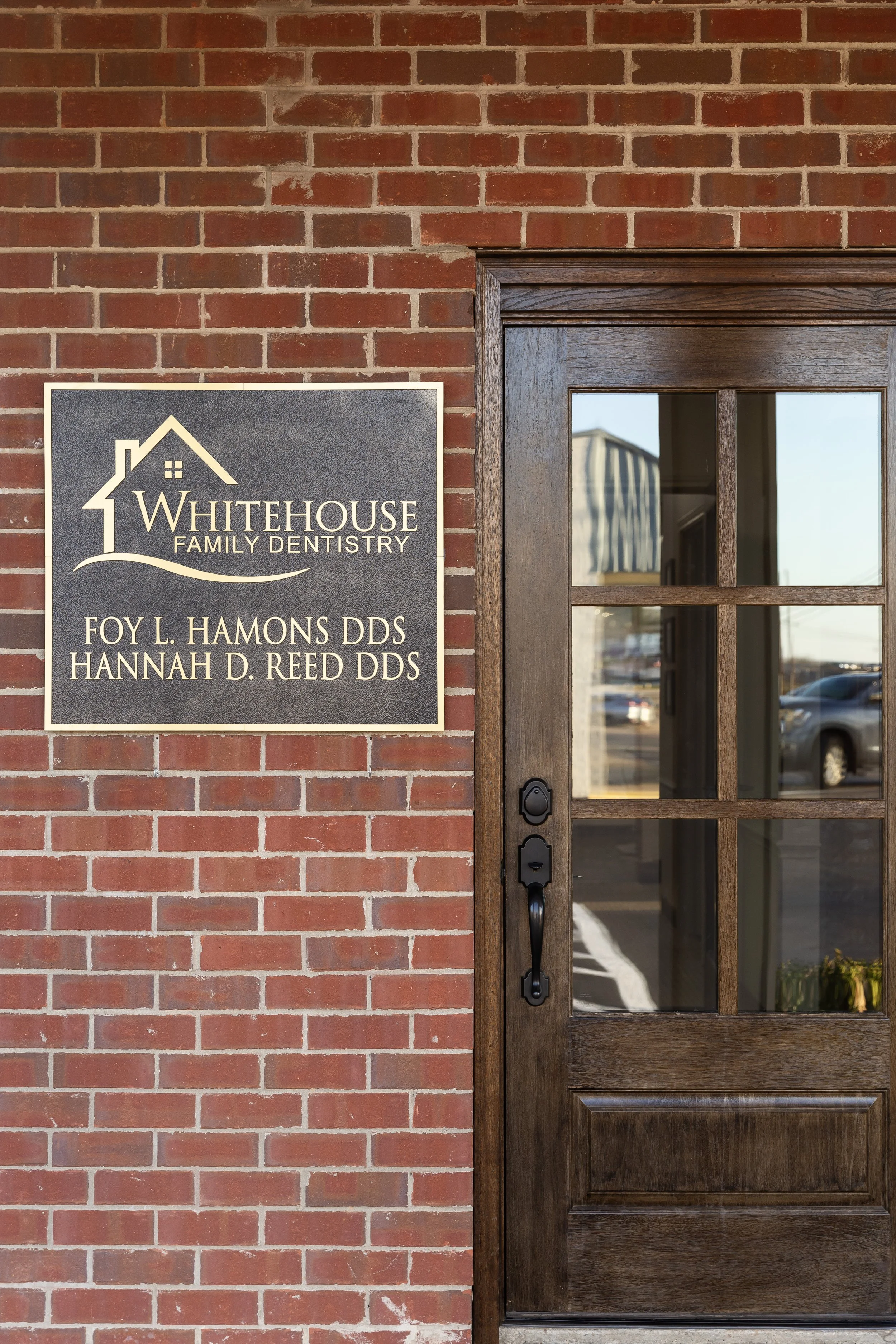 Sign outside a dental office that reads "Whitehouse Family Dentistry" with the names of two dentists, Foy L. Hamons DDS and Hannah D. Reed DDS, next to a wooden door with glass panes.
