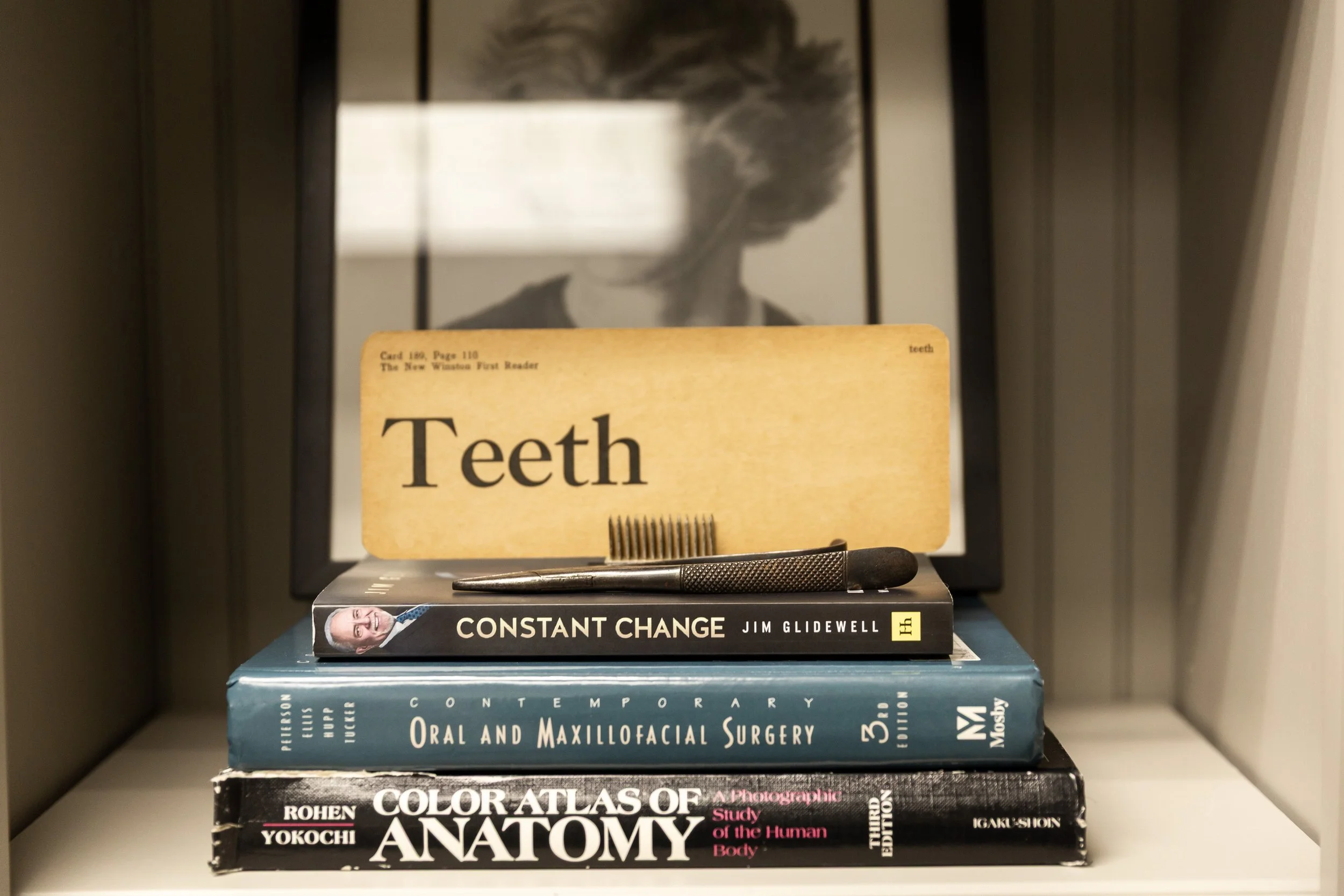 A stack of four medical textbooks and a dental tool on a white shelf. The top book is titled "Constant Change" by Jim Glidewell. The second book is titled "Oral and Maxillofacial Surgery" by Philip J. Strajhar and Vu Nguyen. The third book is titled "Color Atlas of Anatomy" by Rohen Yokochi. The fourth book is titled "Photographic Study of the Human Body." A yellow card with the word "Teeth" stands behind the books, with a hairbrush in front of it. A framed black-and-white photo of someone's head with hair covering the face is in the background.