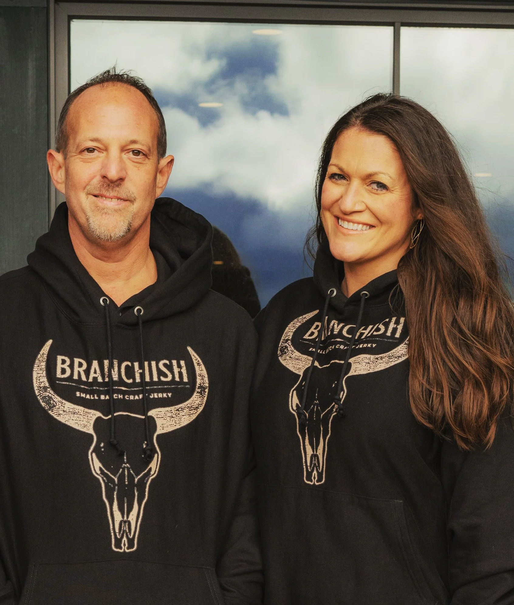 A man and a woman wearing matching black hoodies with a buffalo skull logo and the text 'BRANCHISH Small Batch Craft Jerky' standing in front of a window with a cloudy sky reflection.