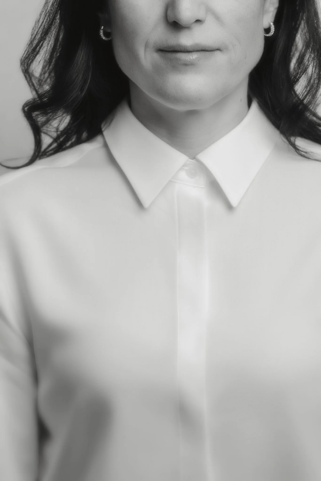 Photo en noir et blanc d'une femme de face, portant une chemise blanche avec col. Seules la bouche, le nez, les oreilles avec boucles d'oreilles et une partie des cheveux sont visibles.