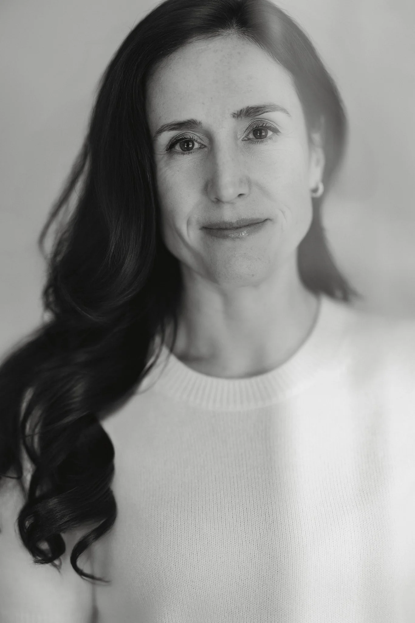 Un portrait en noir et blanc d'une femme avec de longs cheveux bouclés, portant un pull clair et de petites boucles d'oreilles, souriant légèrement.