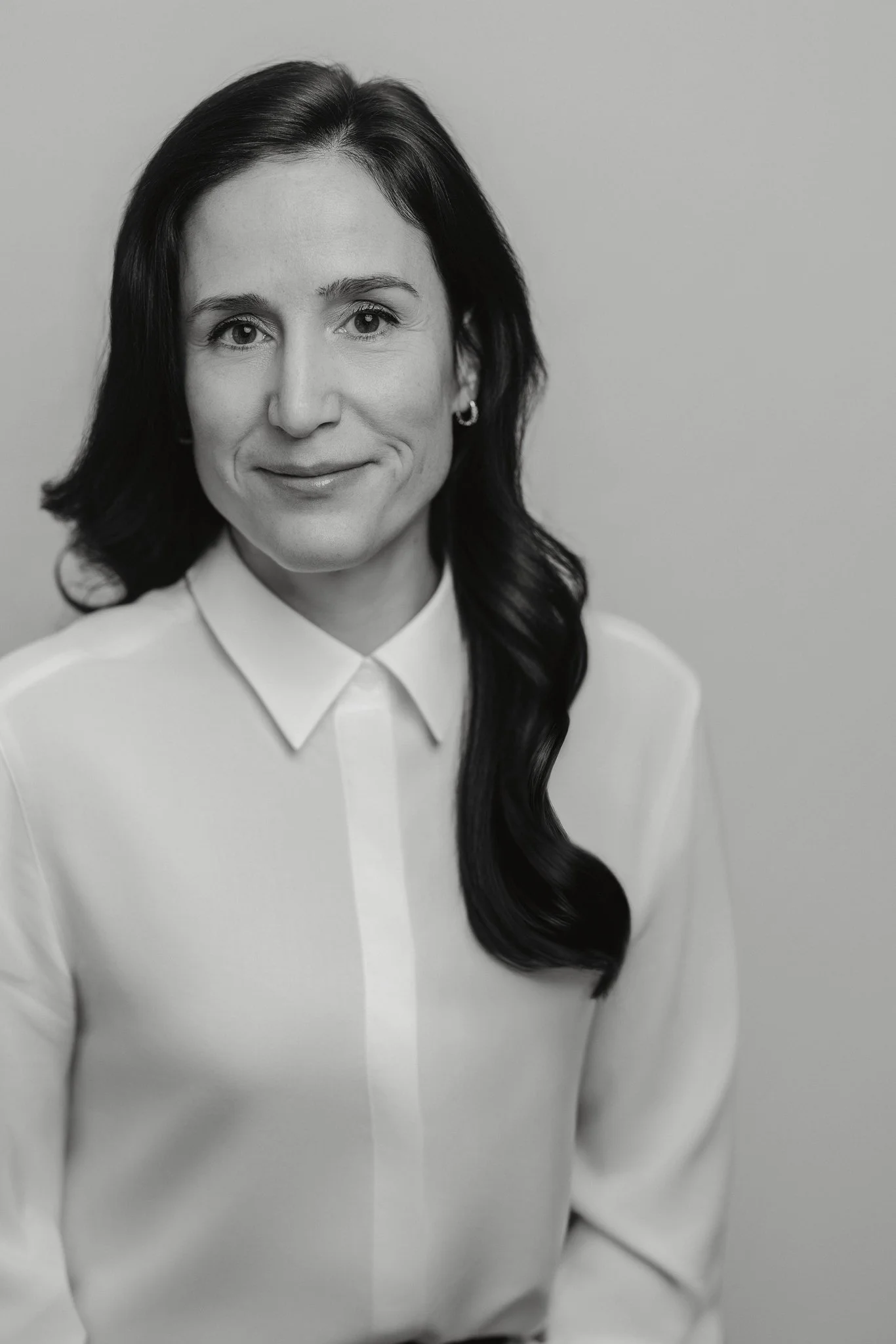 Portrait en noir et blanc d'une femme avec des cheveux foncés, portant une chemise blanche avec col, souriant légèrement, fond uni clair.