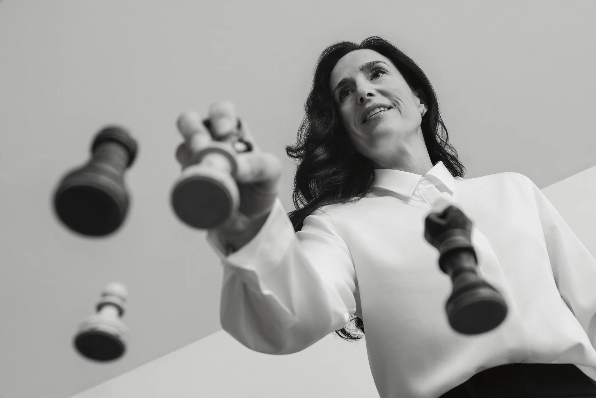 Une femme souriante en gris et blanc, regardant vers le haut, tenant un jeu d'échecs devant elle. L'image est en noir et blanc.