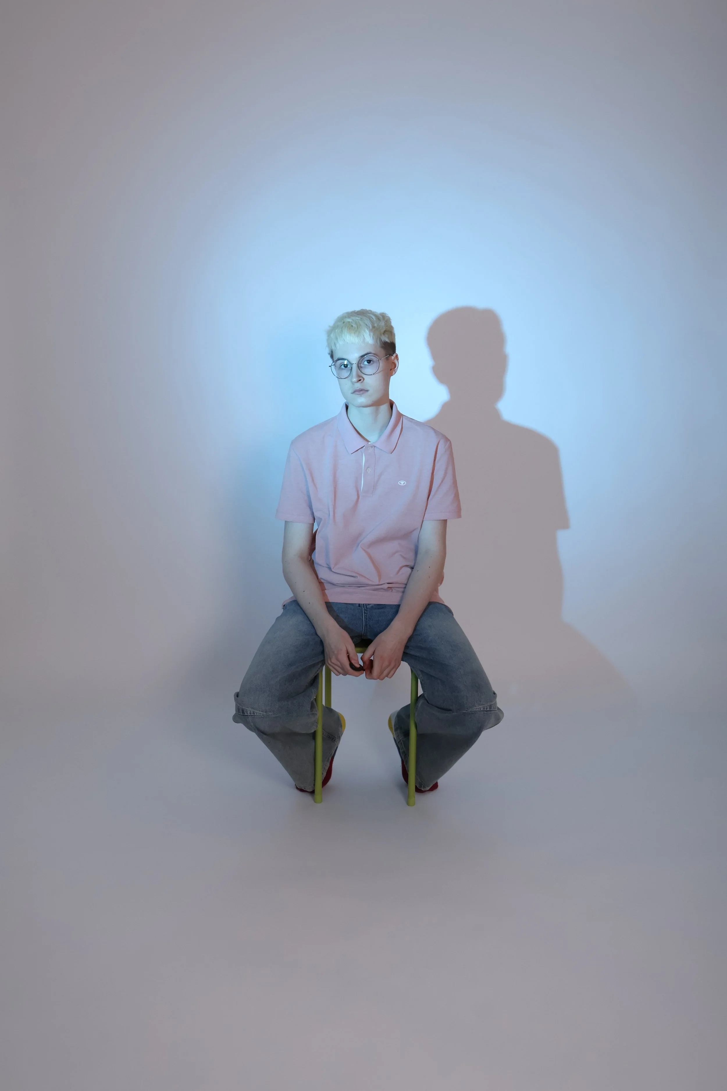 Nuori mies istuu keltaisella jakkaralla, tausta valkoinen, varjo näkyy seinällä.