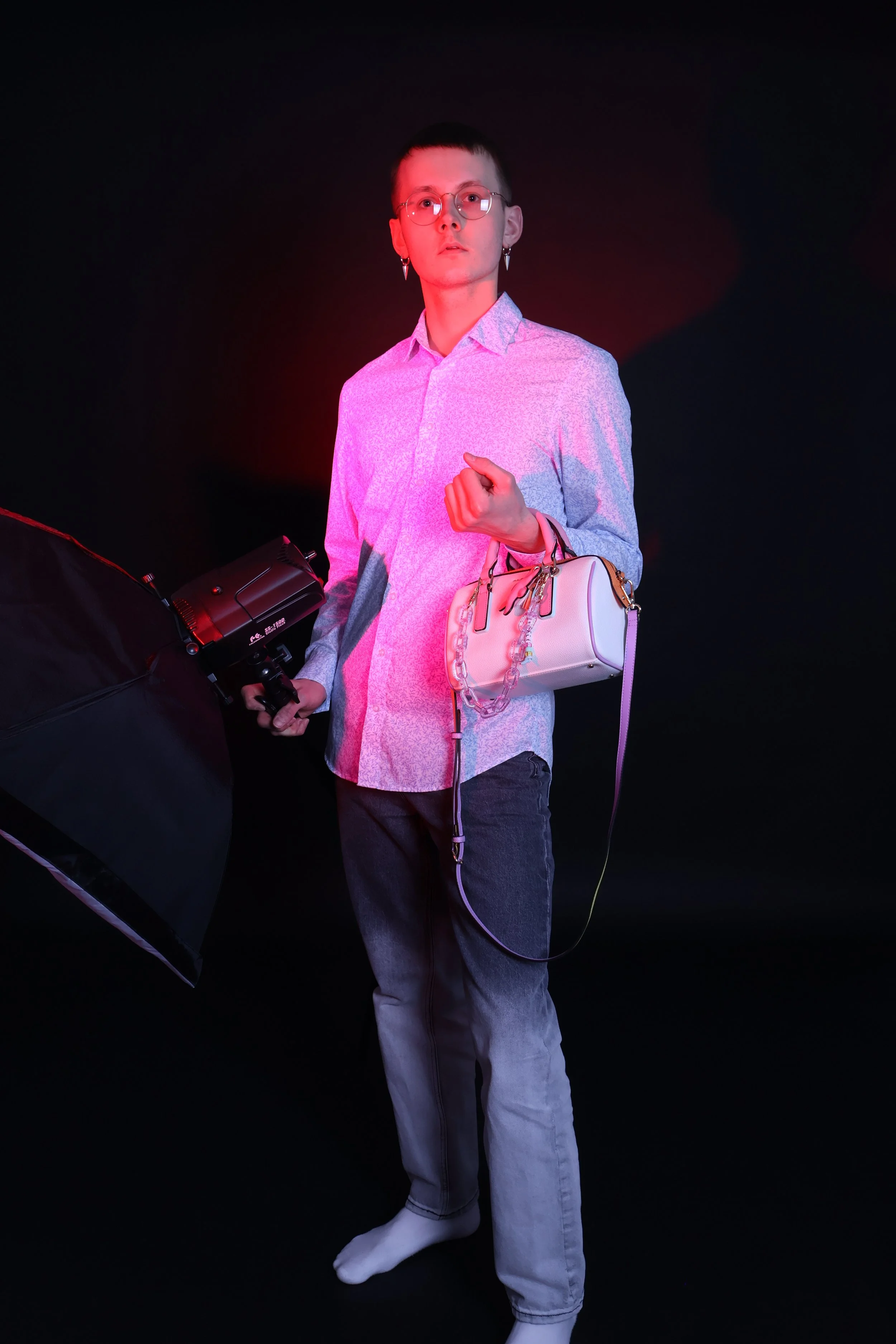 Vauva poseeraa studiossa mustalla taustalla, valaistuna punaisella ja vaaleanpunaisella valolla. Hänellä on muotikuvakoruja ja hän pitää valkoista käsilaukkua ja valkoisia saappaita.