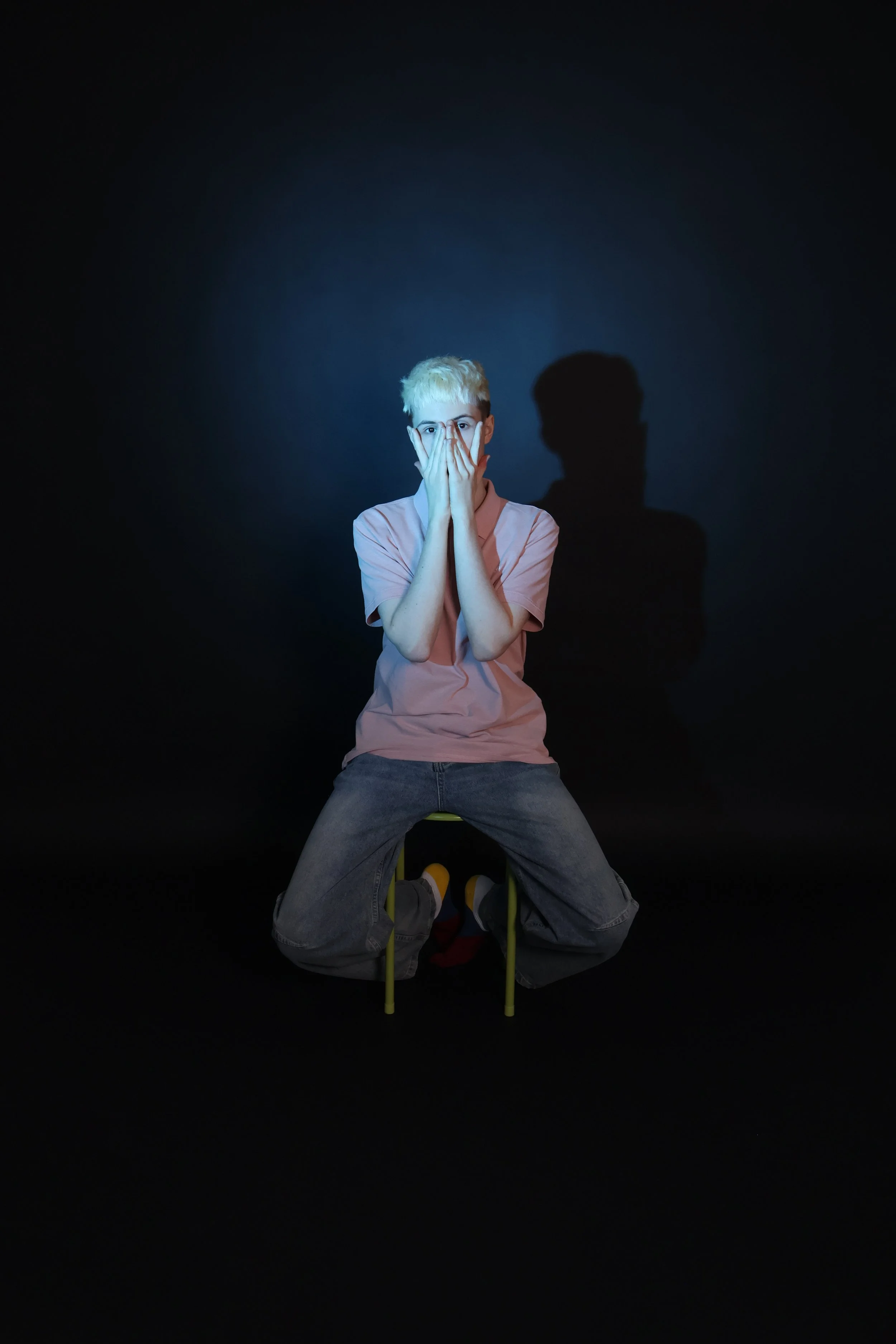 Nuori mies istuu jakkaran päällä ja peittää kasvonsa kädillään pimeässä, taustalla varjo hänestä näkyy seinällä.