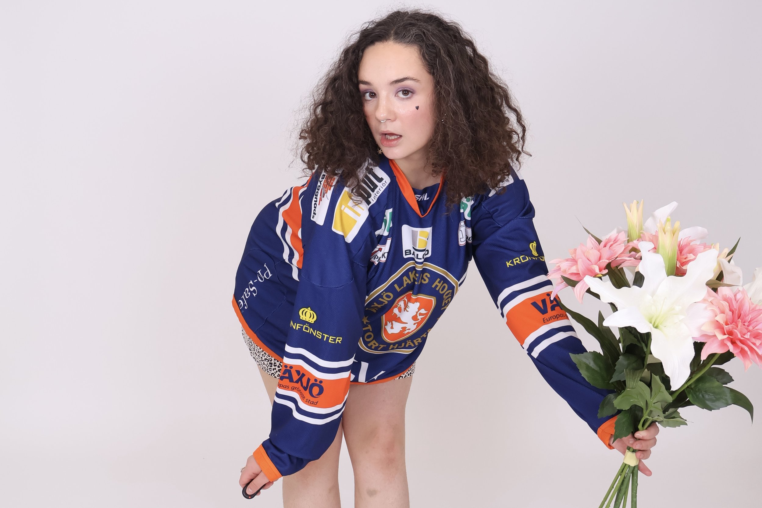 Nuori nainen in hockey-paita esittäytyy kameralle, nojaa yhdellä jalallaan ja pitää kädessään suurta valko- ja vaaleanpunaisä kukkakimppua.