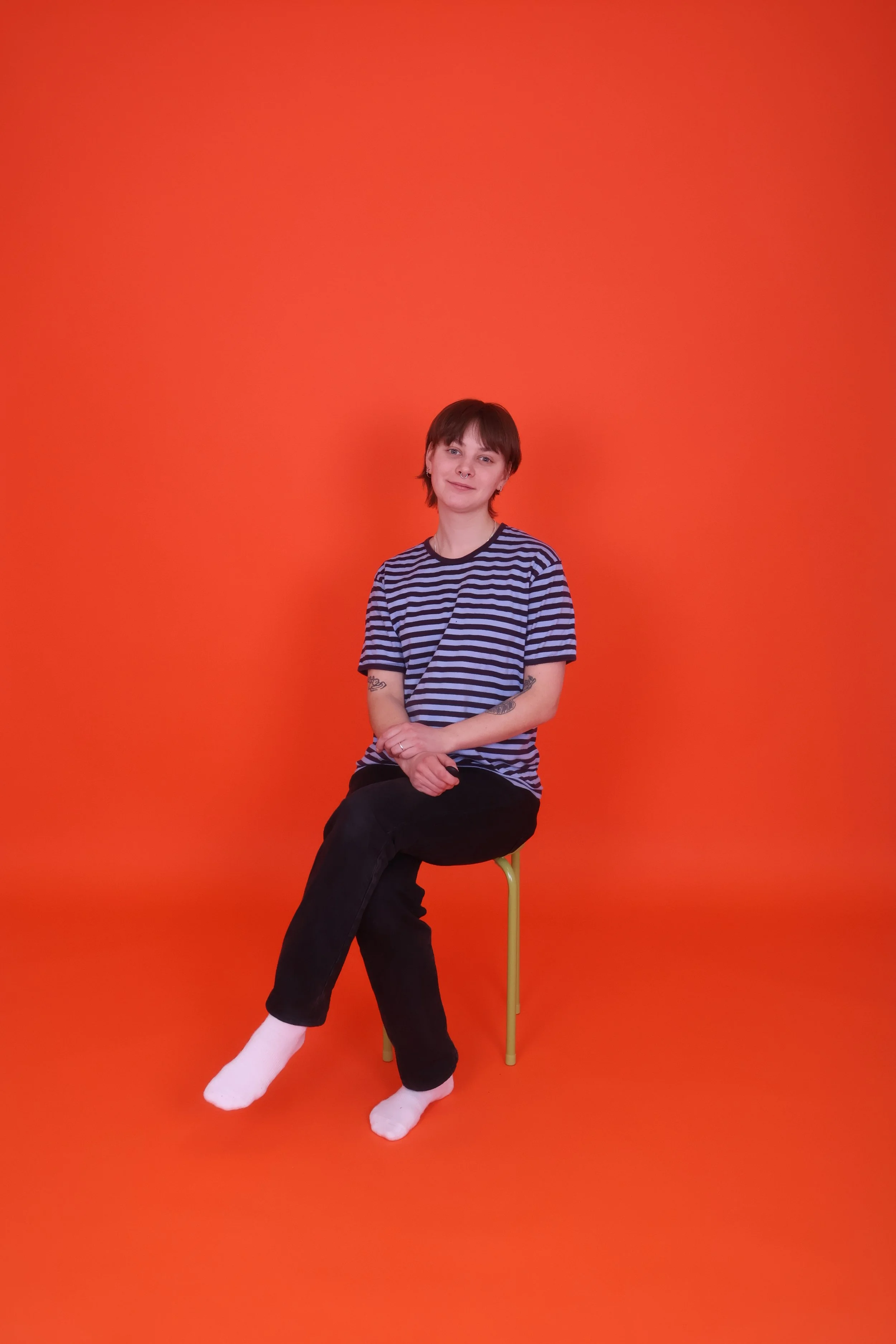 Nuori nainen istuu tuolilla oranssilla taustalla, pukeutuu raidalliseen t-paitaan ja mustiin farkkuihin, ja hänellä on lyhyt ruskea tukka.