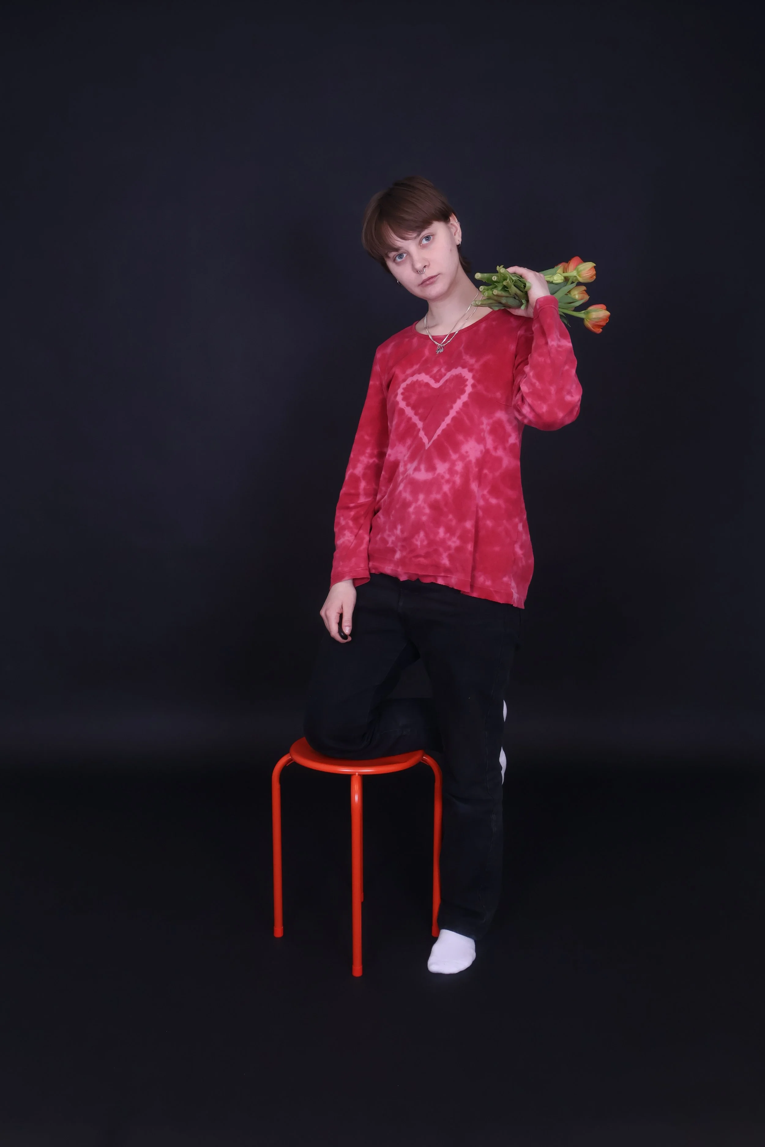 Nuori nainen istuu punaisella jakkaralla mustan taustan edessä, pitää kädessään kimpun tulppaanin kukkia ja katsoo kameraan.