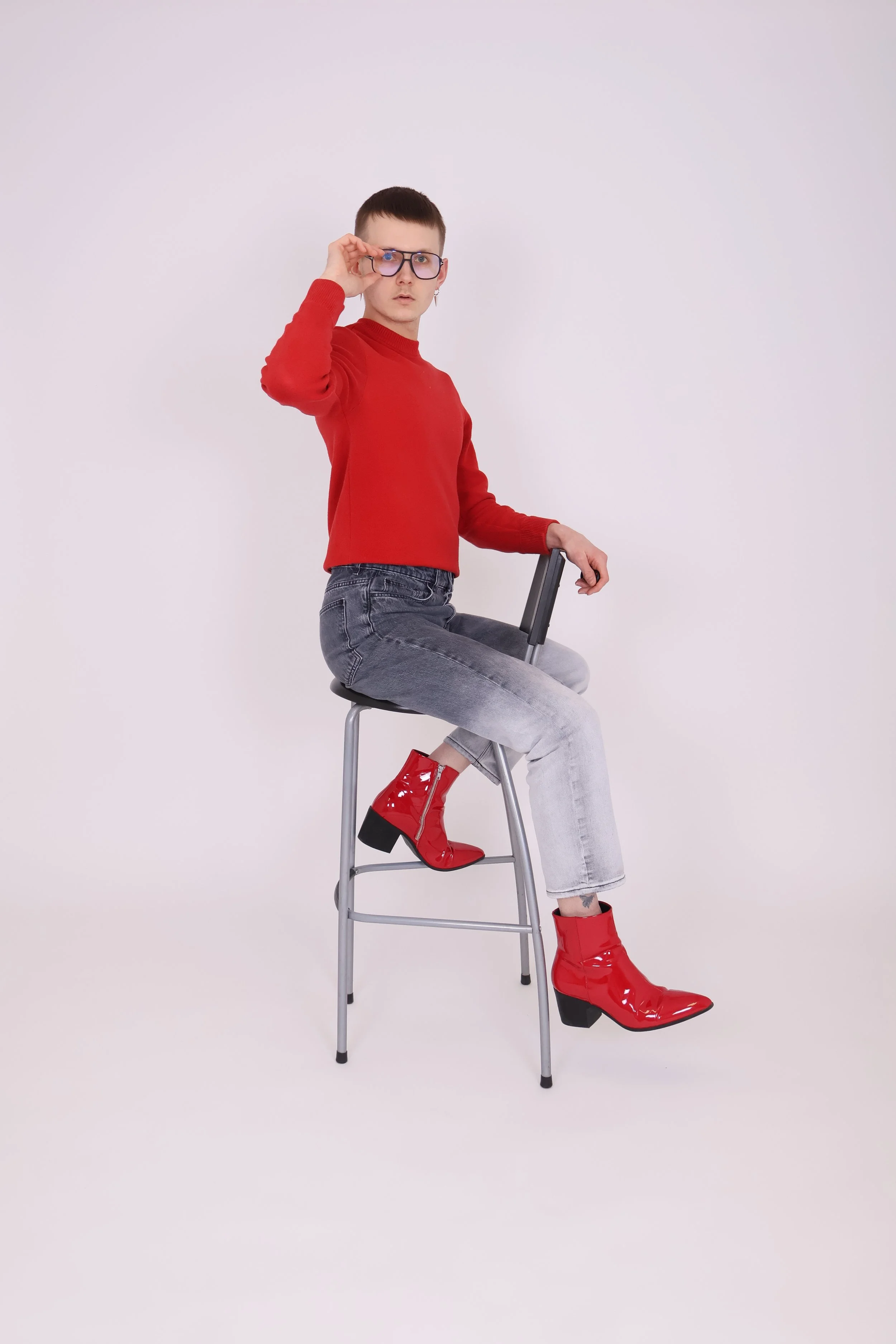 Nuori nainen istuu tuolilla ja katsoo kohti kameraa, käyttää suuria linssejä ja punaista neuletta, hänellä on vaaleat farkut ja kirkkaan punaiset korkokenkäsaappaat.