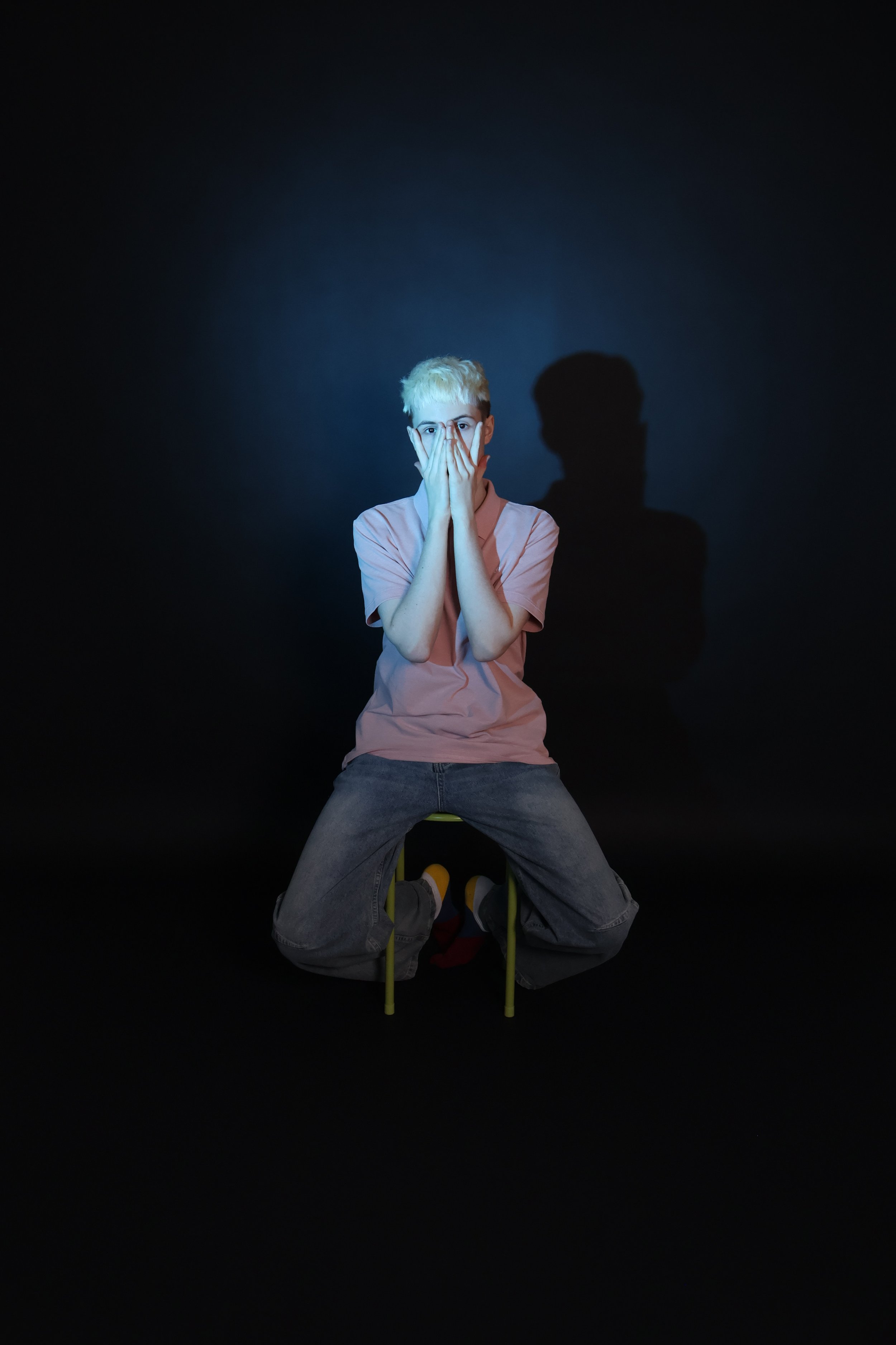 Nuori mies istuu pienellä keltaisella tuolilla pimeässä, valon osuessa häneen ja jättää mustan taustan. Hän peittää suunsa ja nenänsä käsillään ja katsoo suoraan kameraan.