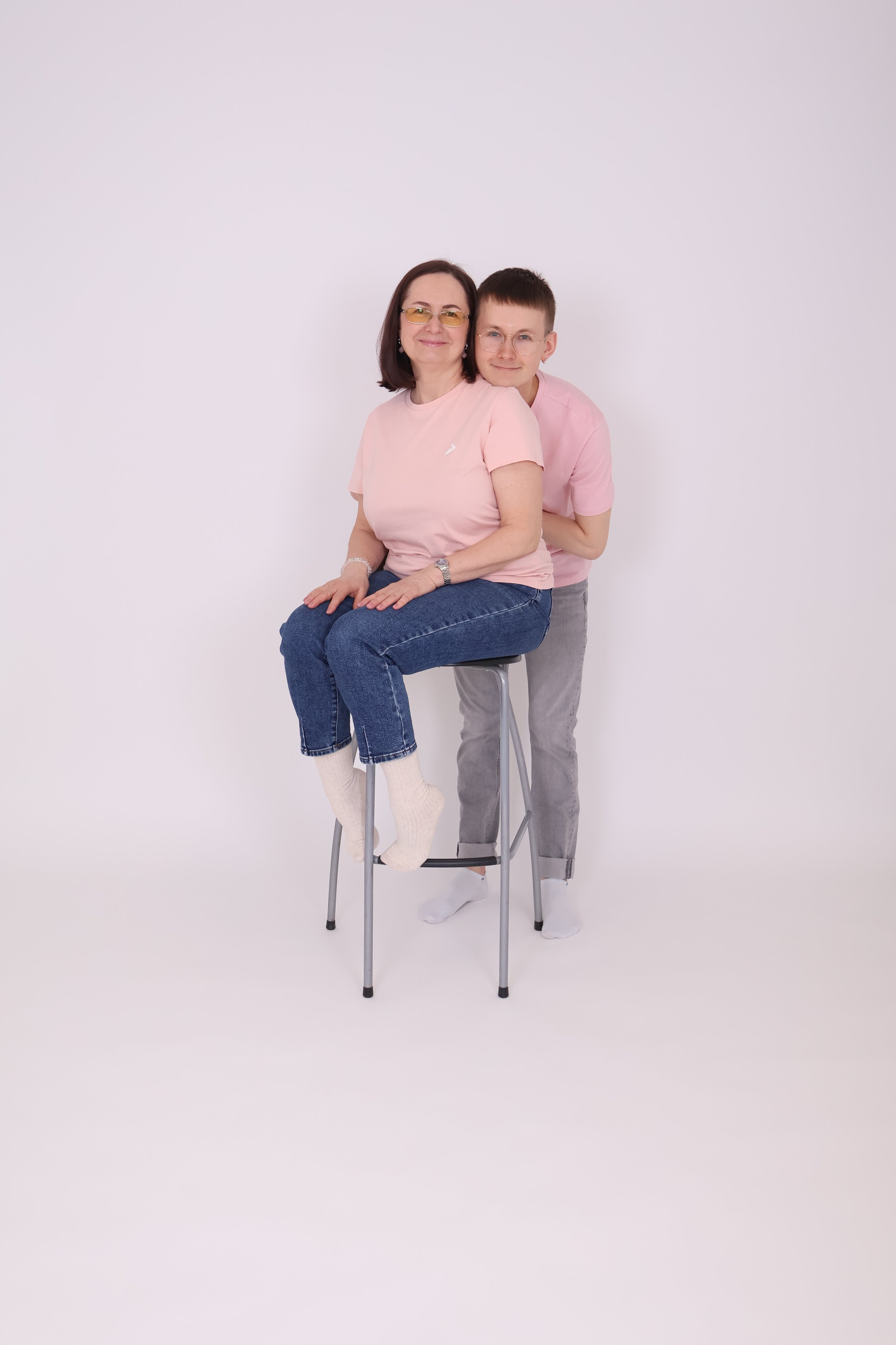 Kuva kahdesta ihmisestä, nainen istuu metallijalustaisella jakkaralla ja nuori mies seisoo hänen takanaan, molemmat hymyilevät ja katselevat kameraa, tausta valkoinen.