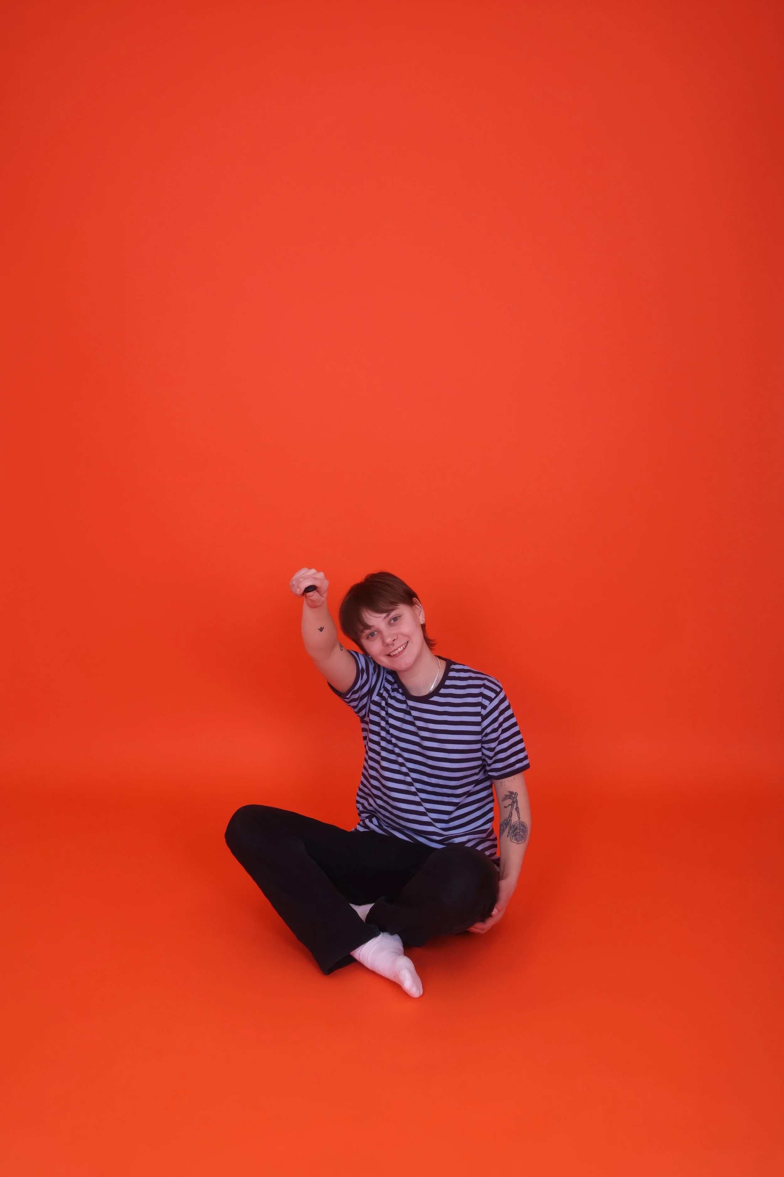 Nuori nainen istuu lattialla, takana tasainen oranssi tausta, hymyilee ja näyttää yhden nyrkin.