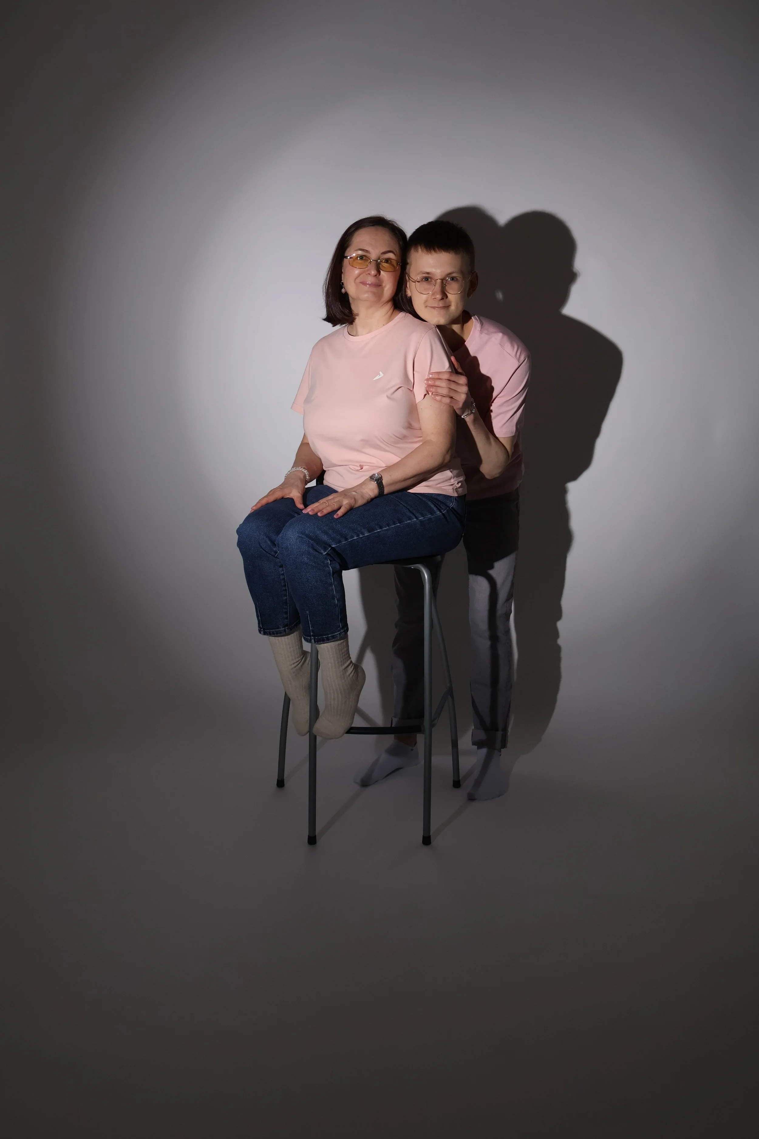 Kaksi ihmistä, äiti ja lapsi, poseeraavat studiokuvassa. Nainen istuu jakkaralla, poika seisoo hänen takanaan ja pitää häntä olkapäästä. Heillä on samanväriset vaatteet ja he katsovat kameraan, taustalla tyhjä valkoinen seinä ja varjot.